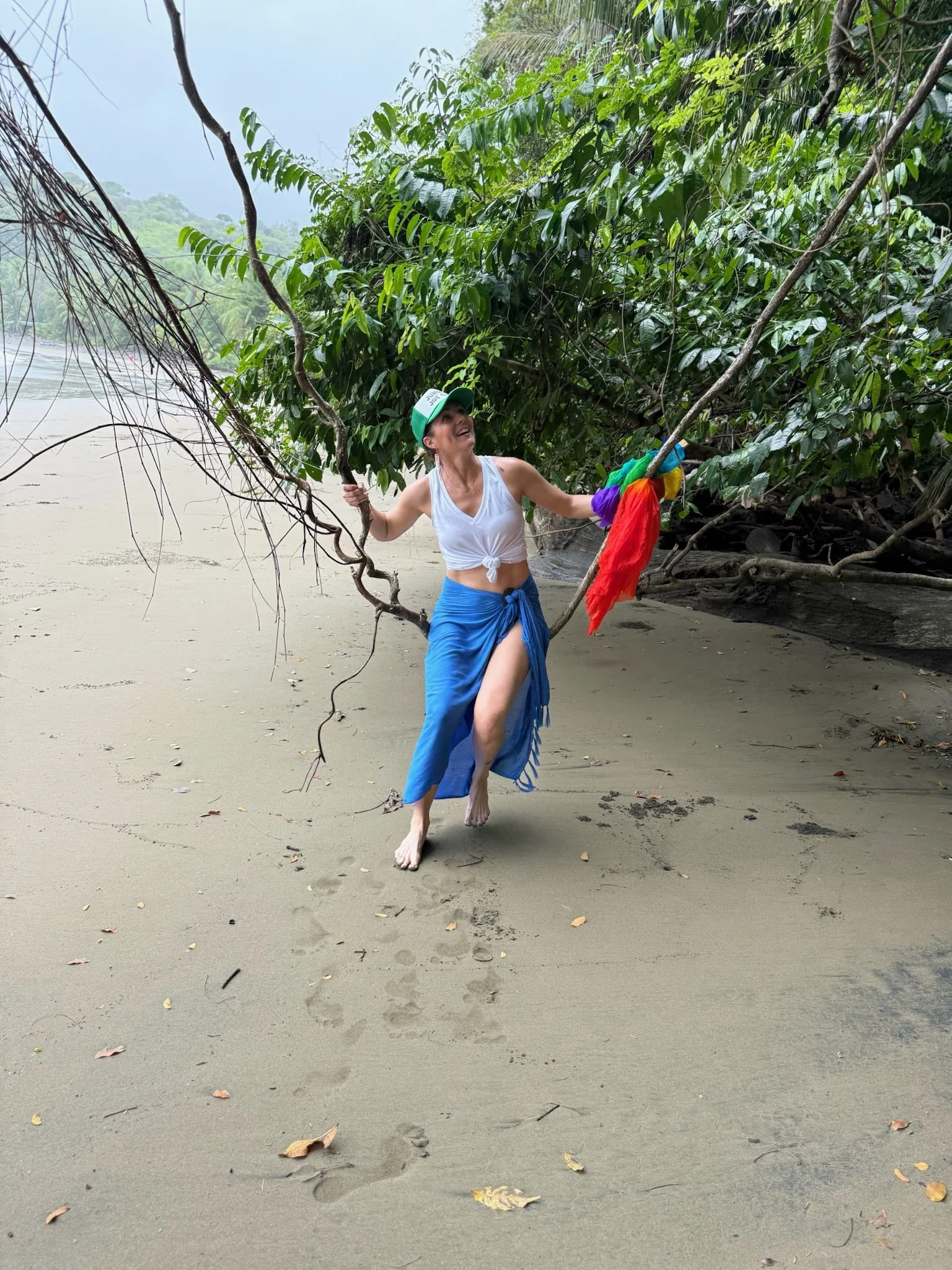 A woman with a white tank top and blue skirt holding onto branches of a tree on a sandy beach with footprints and fallen leaves, with green foliage and trees in the background.