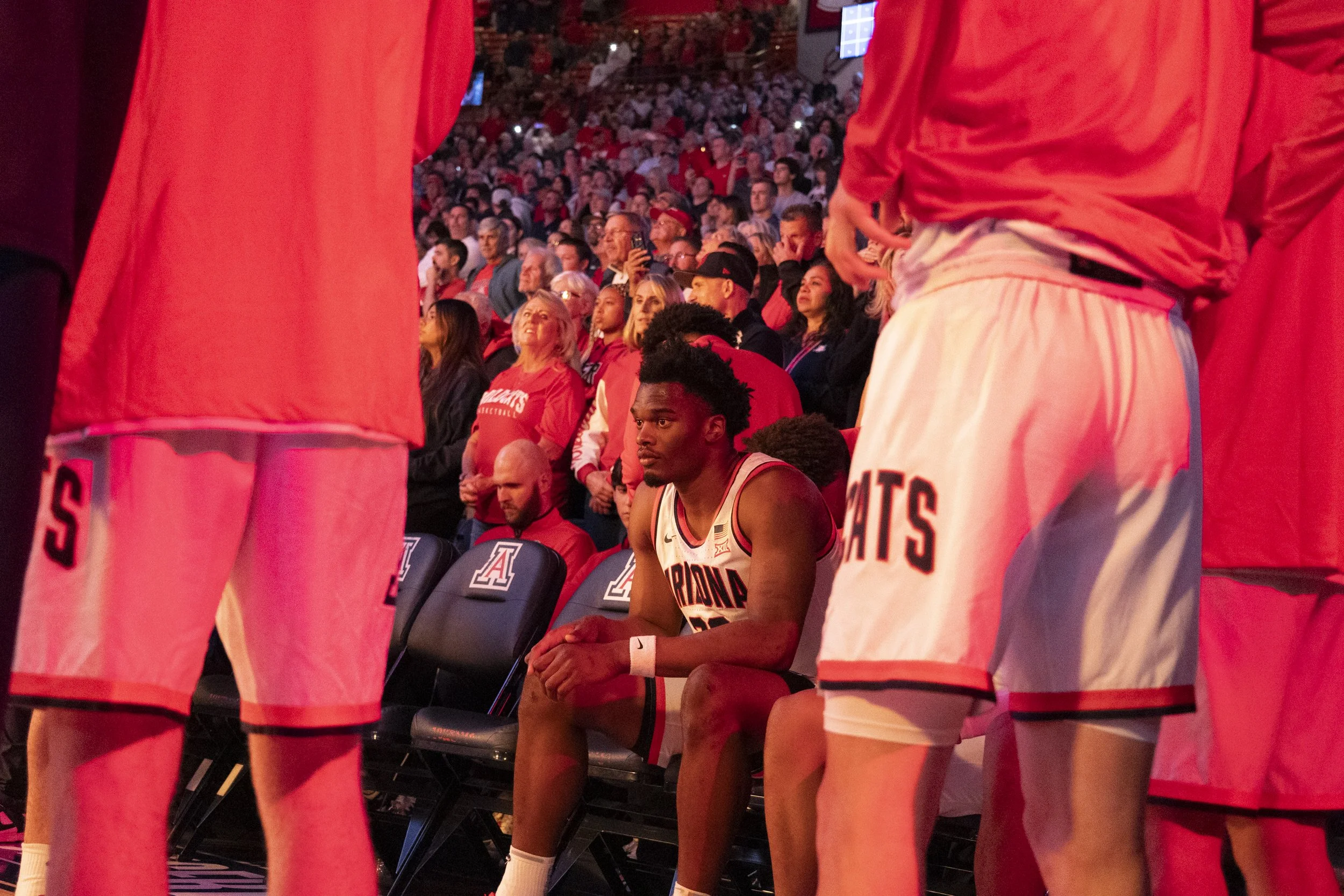 A basketball player sitting on a bench surrounded by teammates during a game, with a large crowd watching in the background.