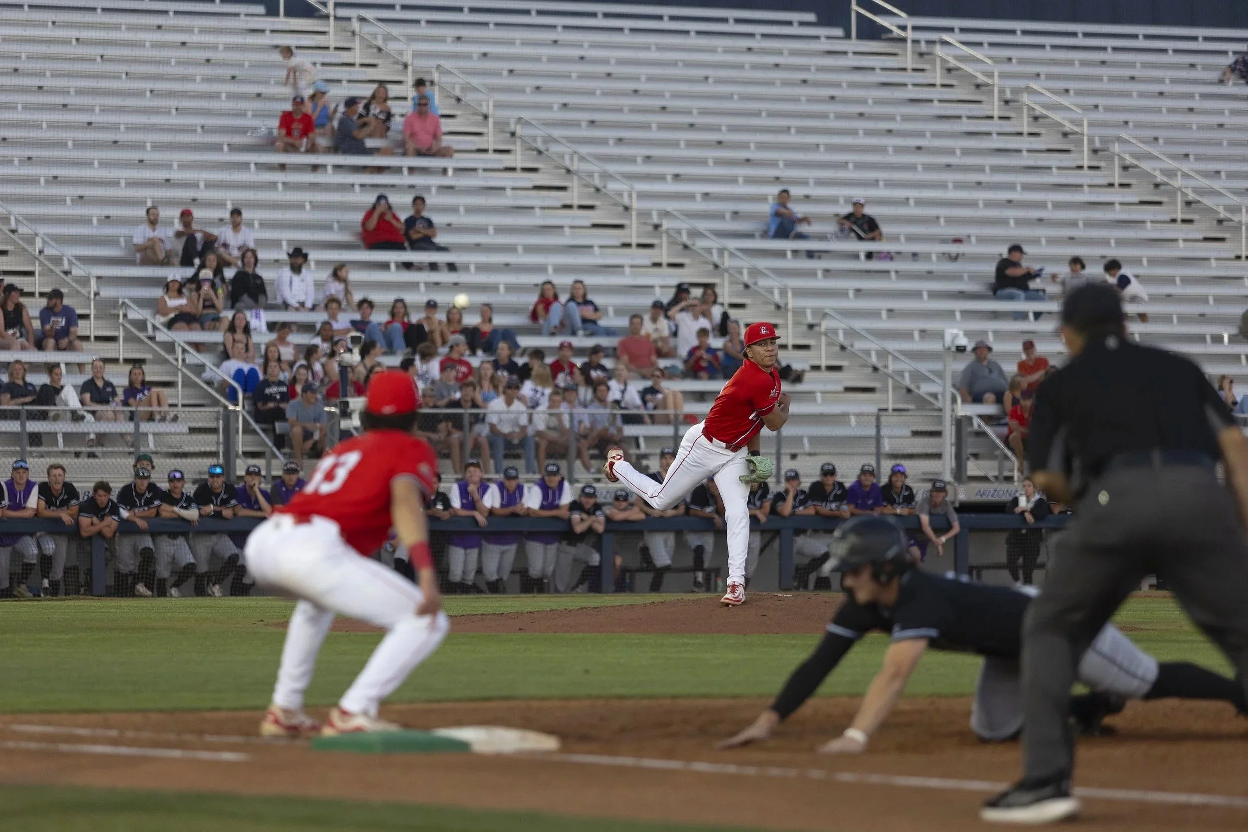A baseball player in a red jersey and white pants running around the bases during a game, with other players, an umpire, and spectators in the background.
