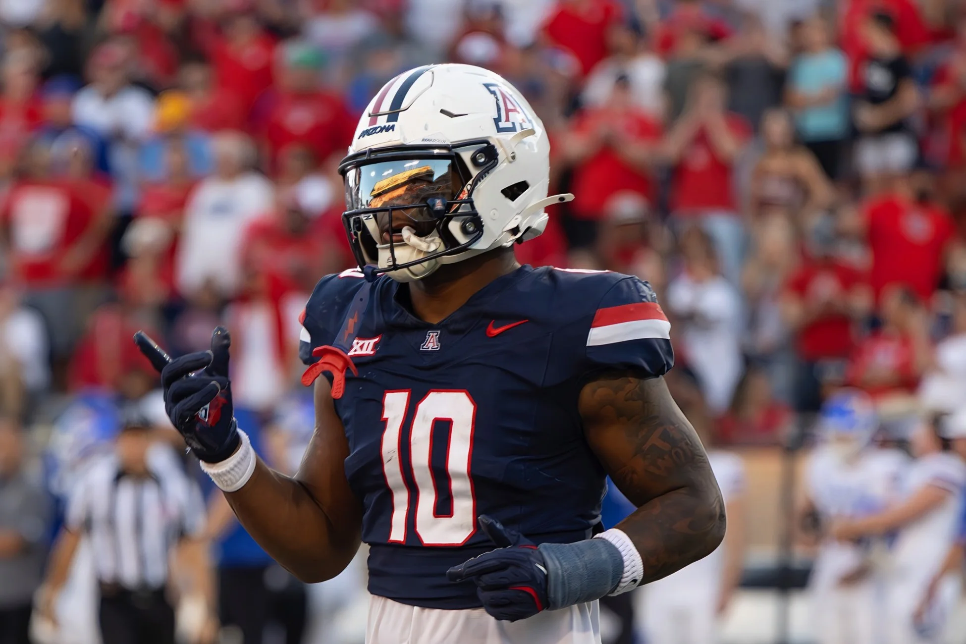 A University of Arizona football player wearing a dark blue jersey with the number 10, white helmet with a logo, and dark gloves, is captured during a game with a crowded stadium in the background.
