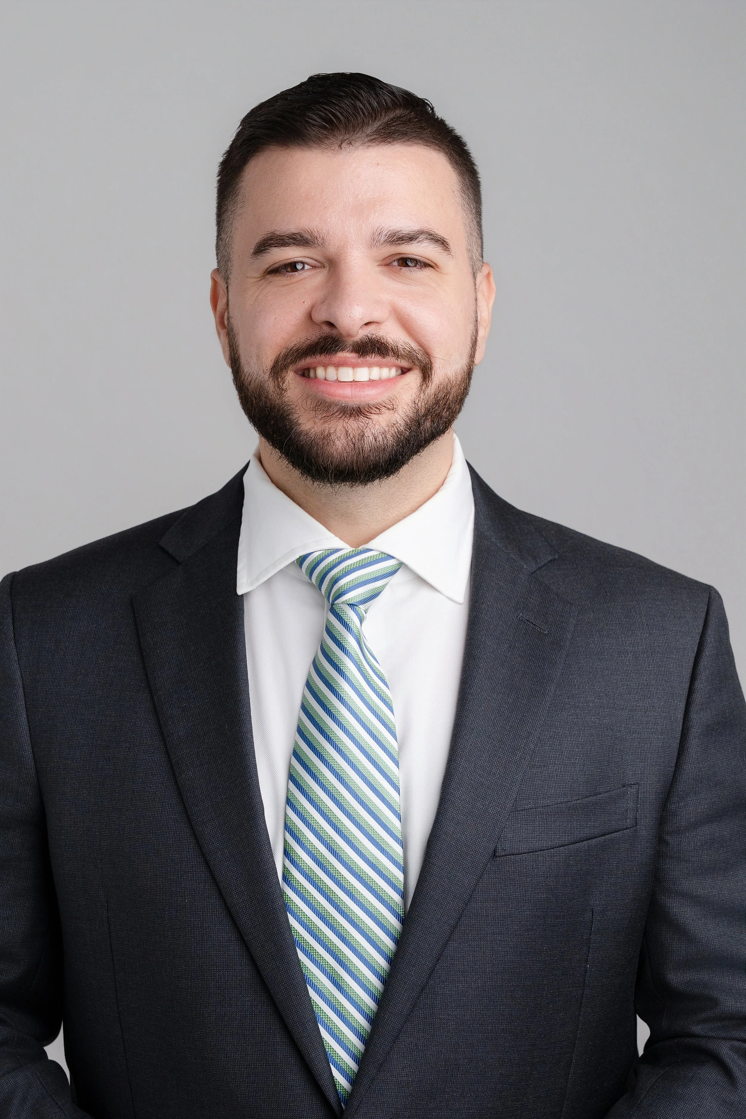 A professional headshot of a smiling man with short dark hair, a beard, wearing a dark suit, white shirt, and a striped green and blue tie, posing against a plain grey background.