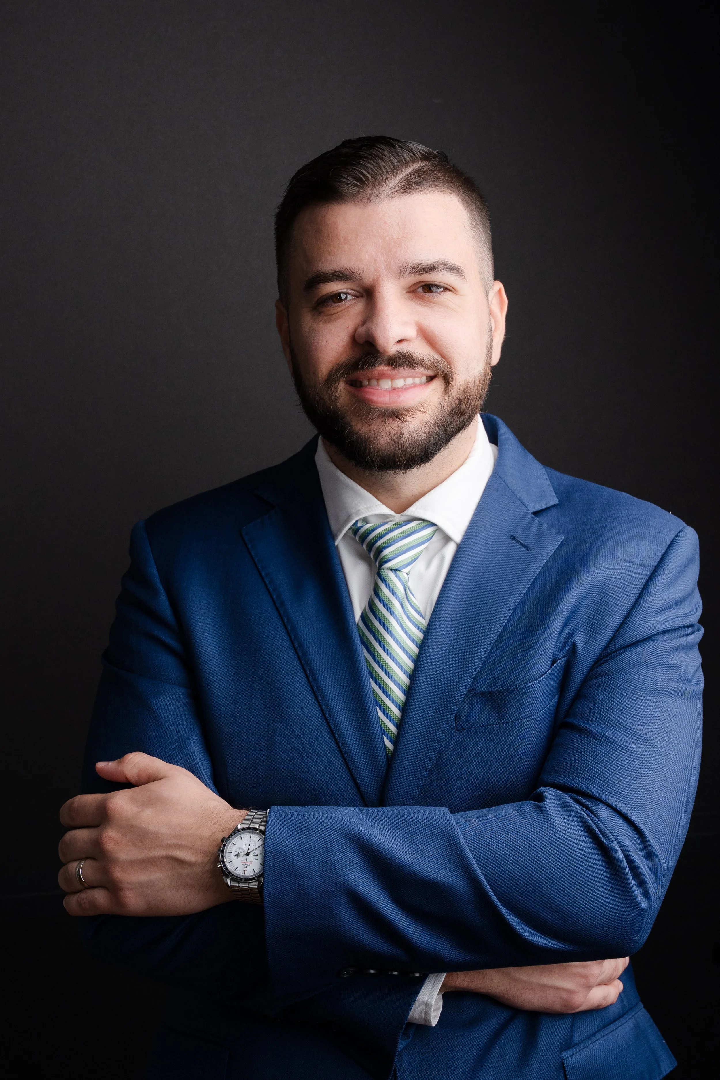 A man in a blue suit with a striped tie smiling, with arms crossed, wearing a wristwatch, omega speedmaster professional, against a dark background.