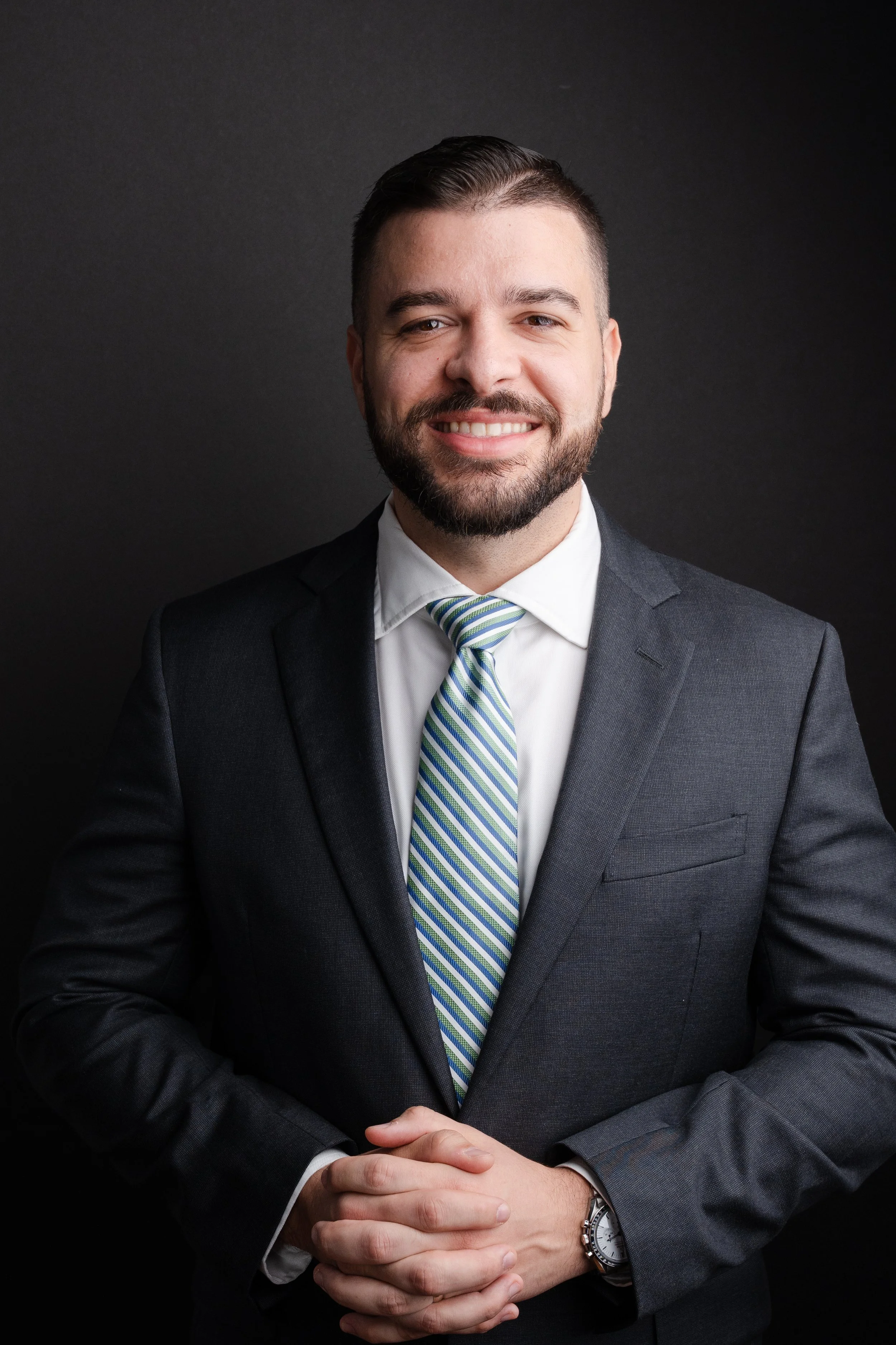 A smiling man in a dark suit with a white shirt and a striped tie, standing against a black background. He has short dark hair and a beard, with his hands clasped in front of him, wearing a silver wristwatch, an omega speedmaster professional.