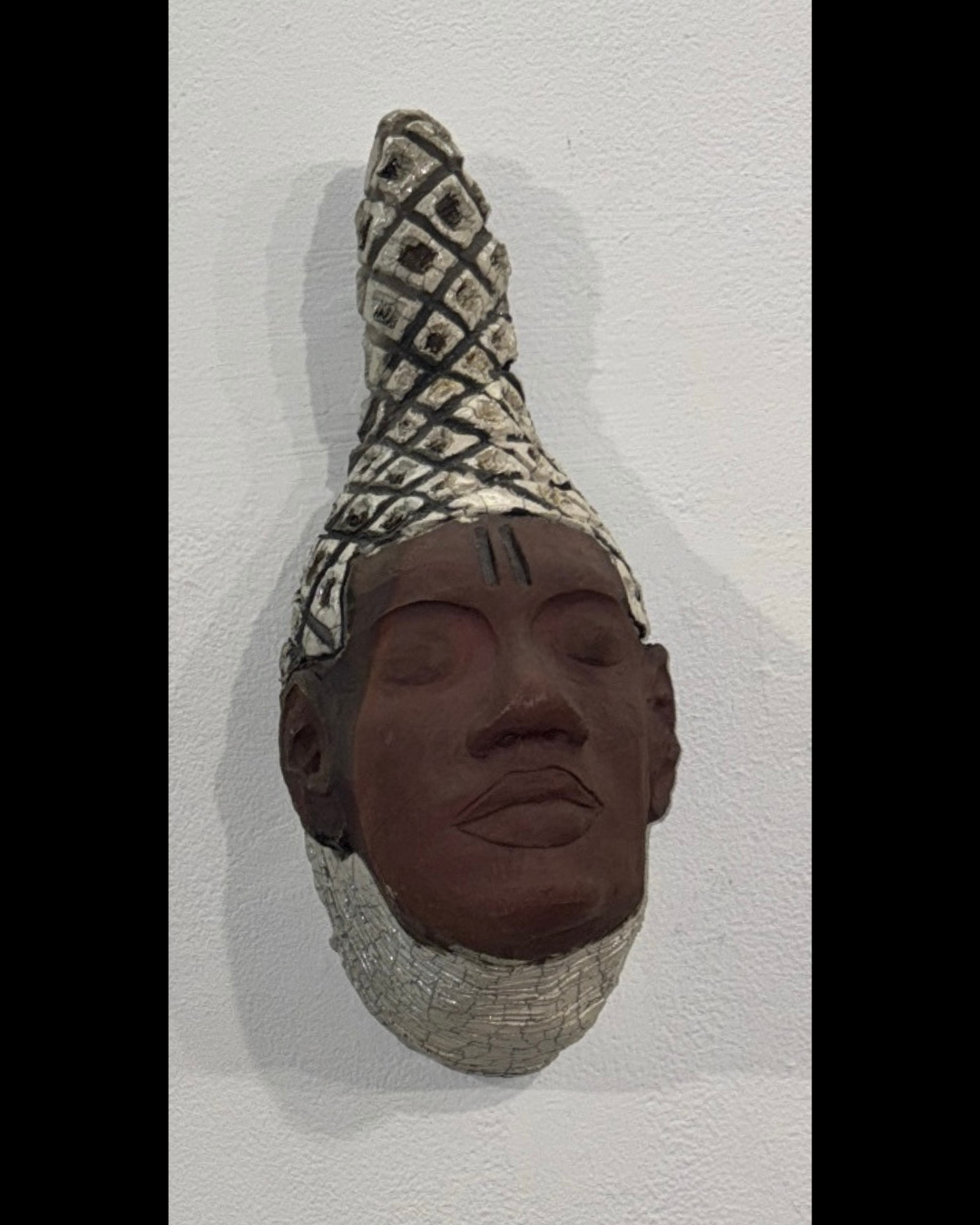 A wall-mounted sculpture of a person's face with closed eyes, dark skin tone, full lips, and a downward-tilted head, wearing a patterned headwrap with black and white geometric designs.