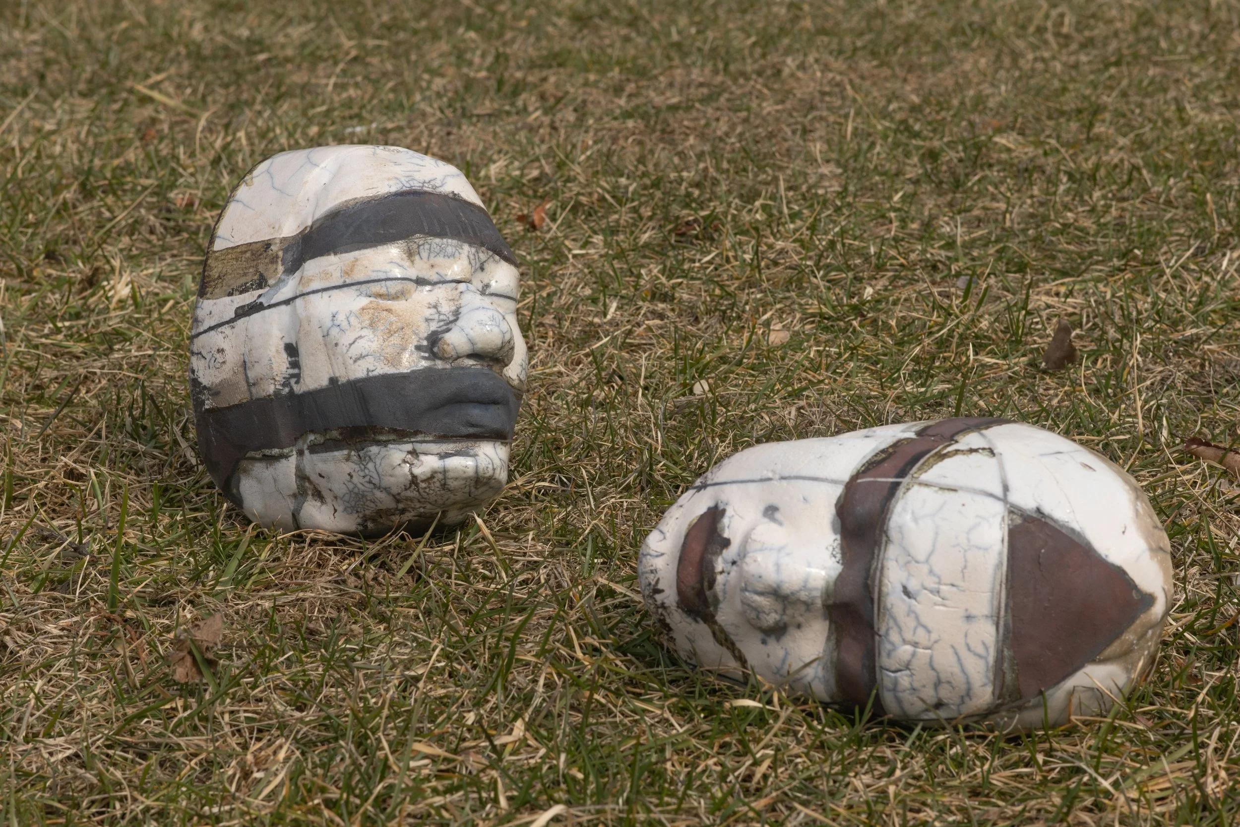 Several cracked and painted skull sculptures lying on dry grass in an outdoor setting.