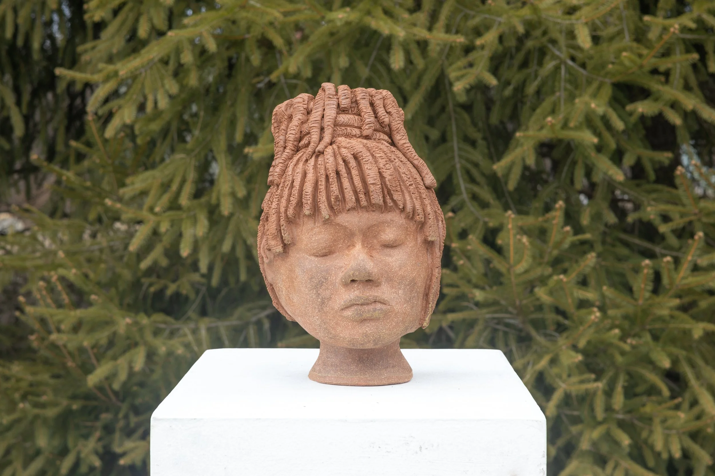 A sculpture of a person's head with closed eyes and dreadlock hairstyle set on a white pedestal in front of green tree branches.