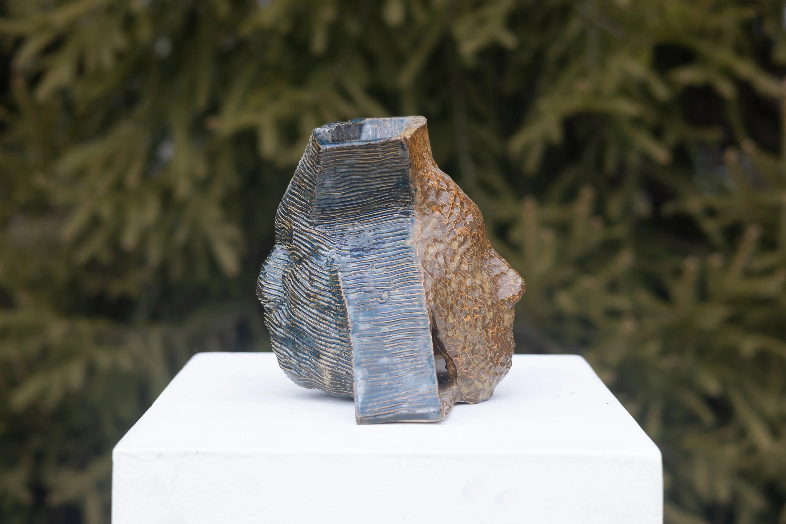 A close-up of an abstract sculpture with textured and layered patterns, displayed outdoors on a white pedestal with greenery in the background.