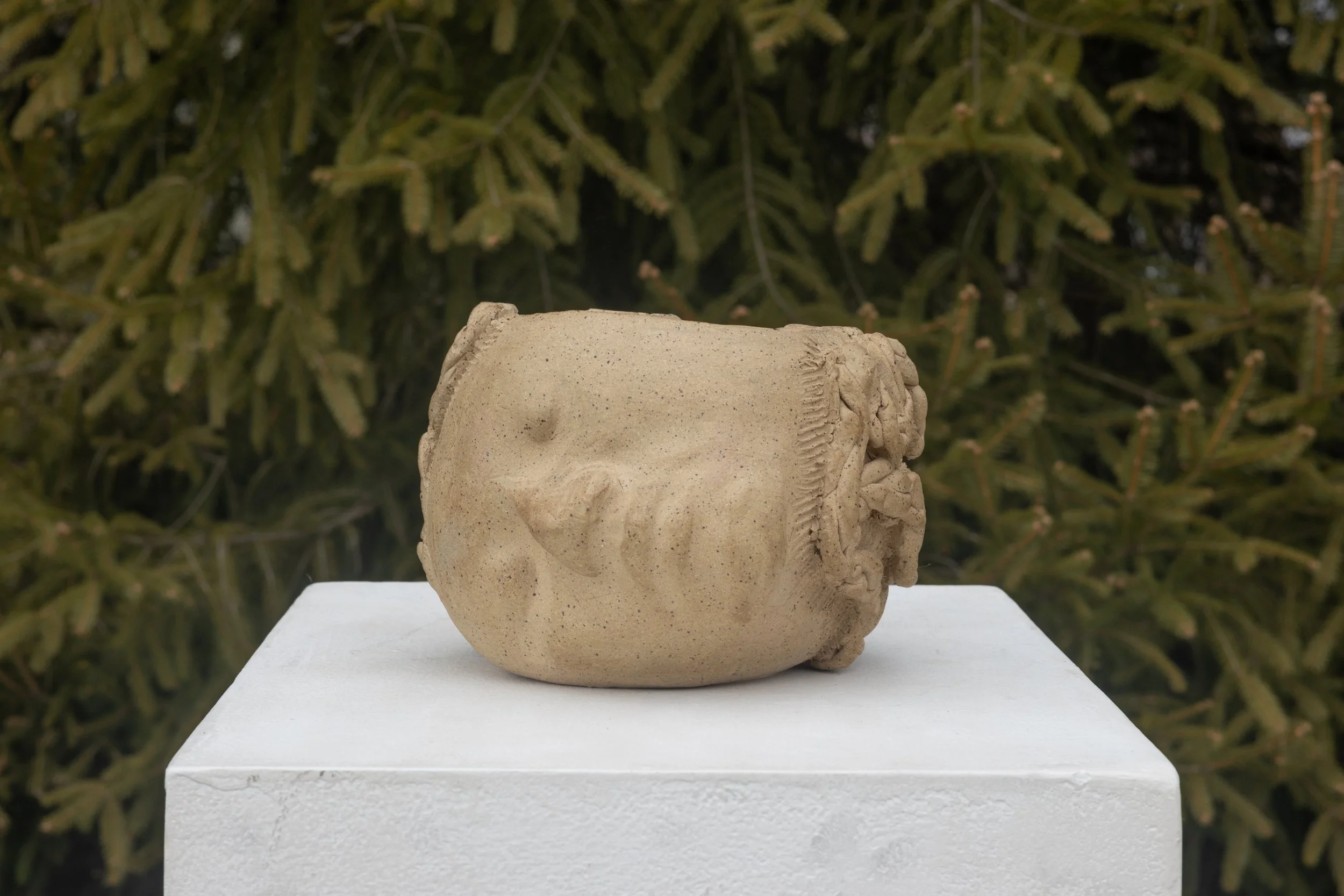 Ancient sandstone sculpture of a human head placed on a white pedestal outdoors with lush green trees in the background.