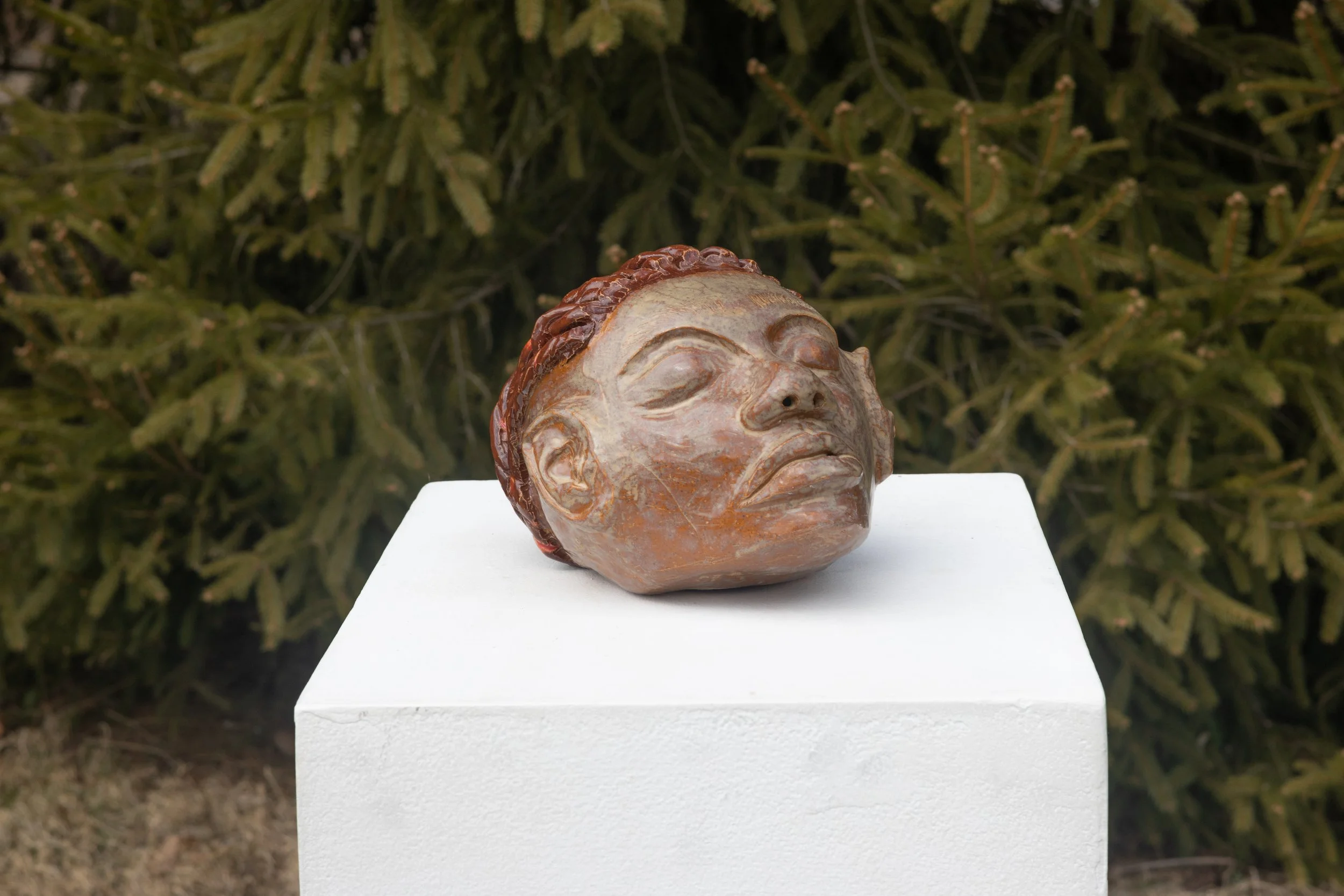 Ceramic sculpture of a human head with closed eyes and lips, placed on a white pedestal outdoors.