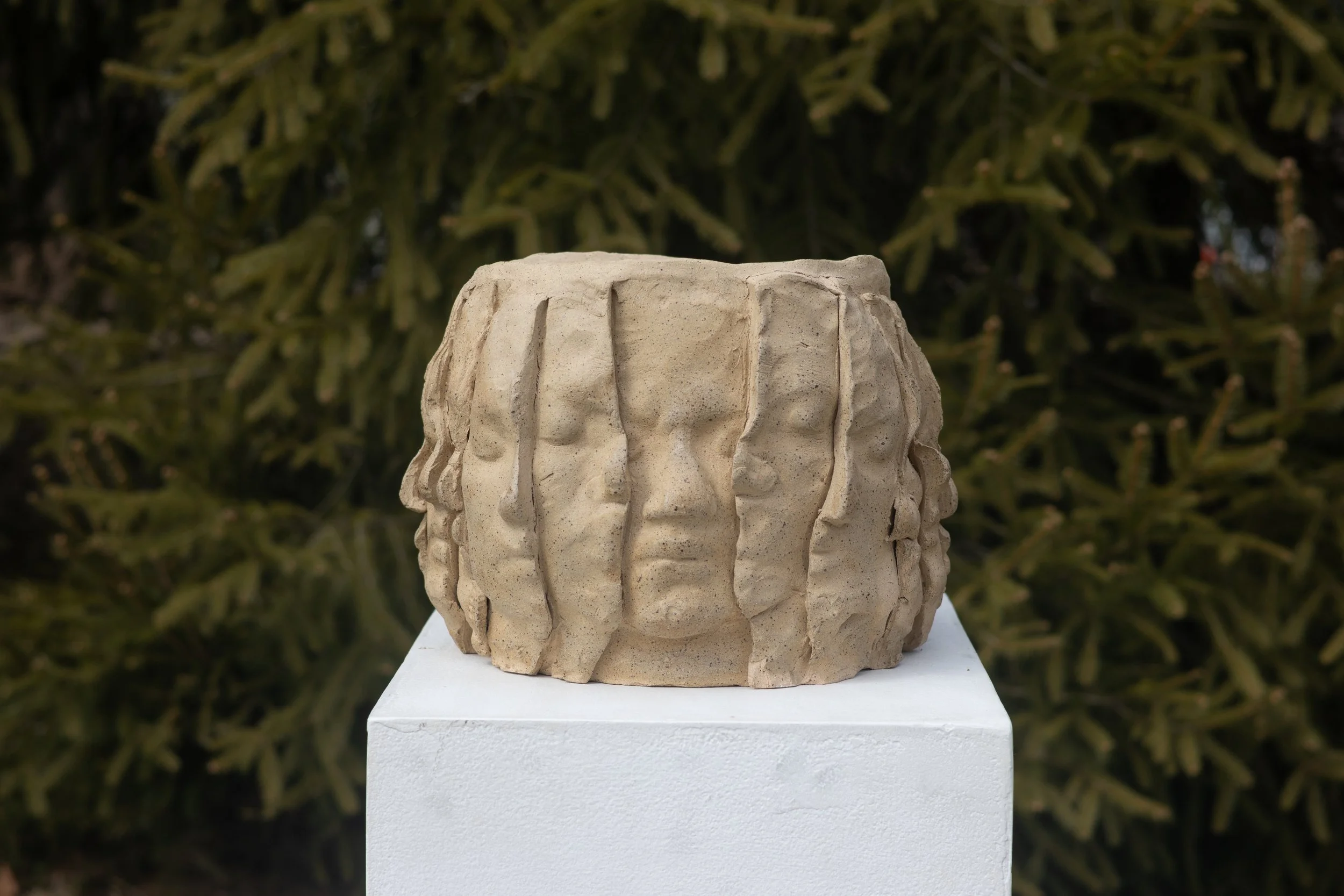 A beige ceramic sculpture of a face with long hair, placed on a white pedestal outdoors with green foliage in the background.