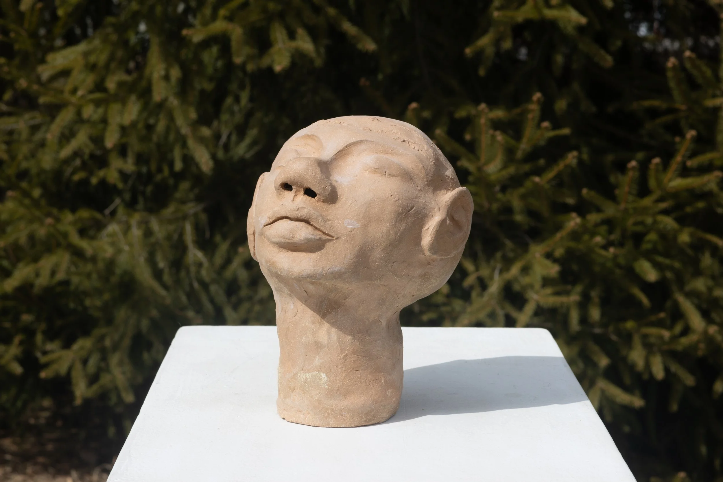 Sculpture of a human head with closed eyes, made of light-colored material, placed on a white pedestal outdoors with greenery in the background.