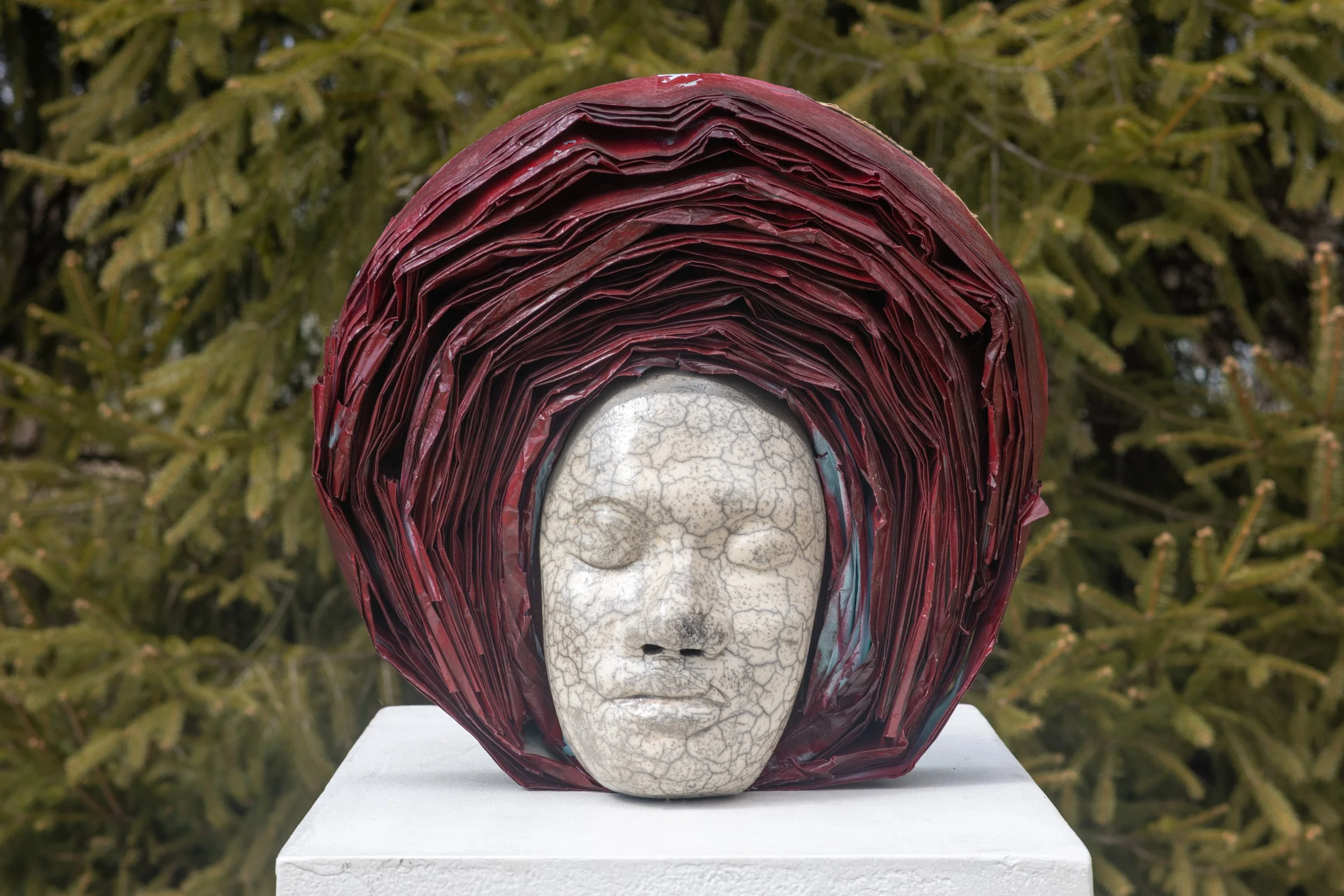 A sculpture featuring a stylized face with a cracked texture, partially covered by a layered red and multicolored material resembling a hood or hair, set outdoors with green foliage in the background.