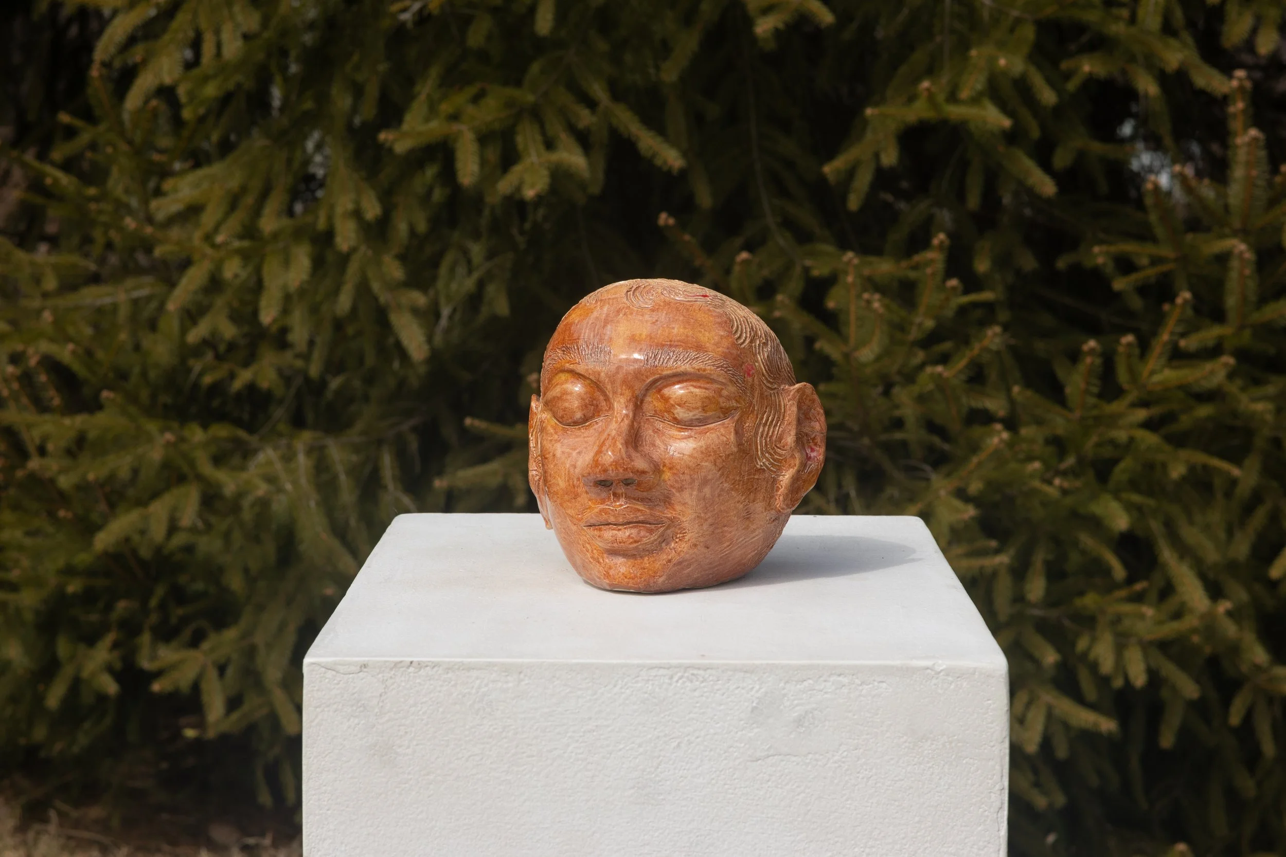 A carved stone sculpture of a person's face with eyes closed, placed on a white pedestal outdoors with green foliage in the background.