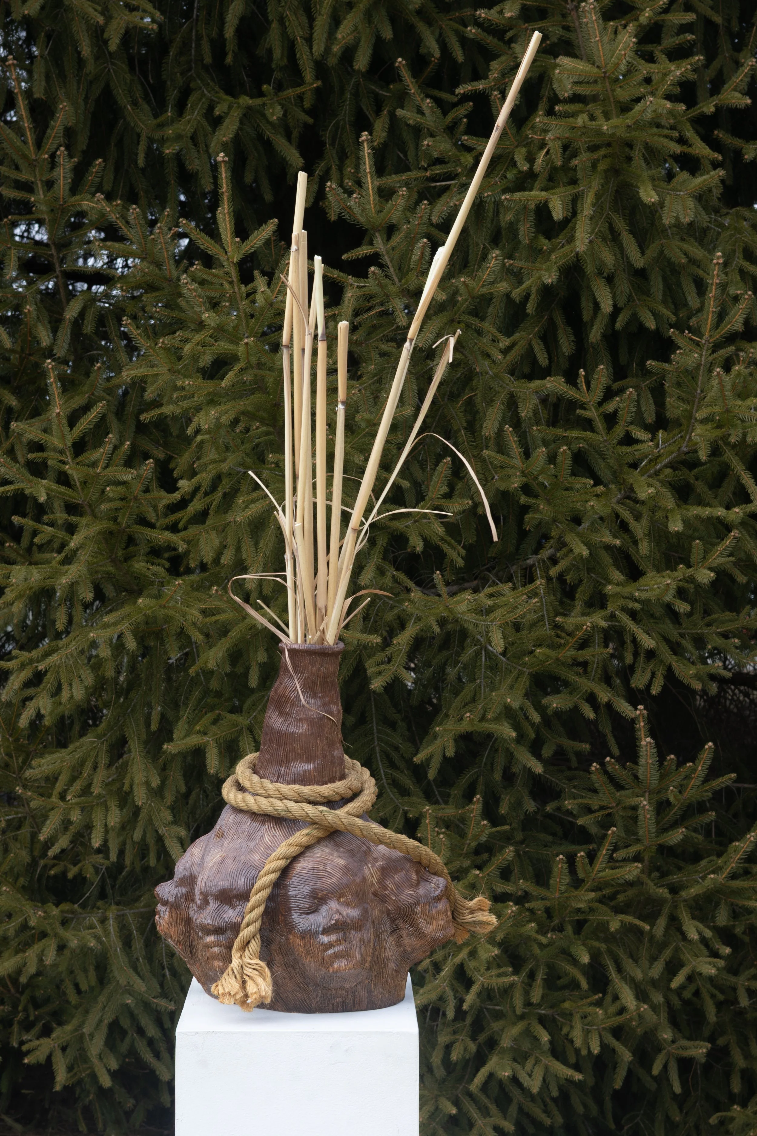 Wooden sculpture resembling a person with a rounded head, adorned with a thick rope, surrounded by thin bamboo sticks, and placed on a white pedestal outdoors, with greenery in the background.
