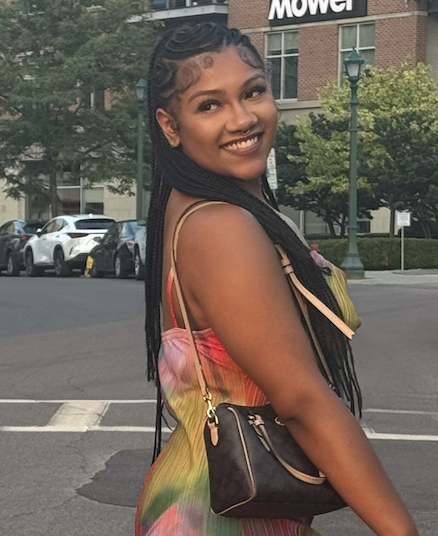 A young woman with long braided hair smiling outdoors on a city street, wearing a colorful dress and holding a black purse.