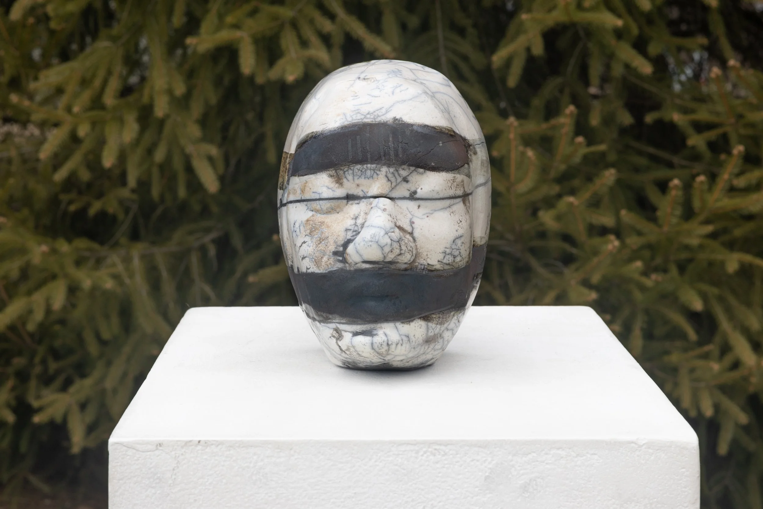 Marble stone sculpture of a face with dark markings, displayed on a white pedestal outdoors against green foliage.