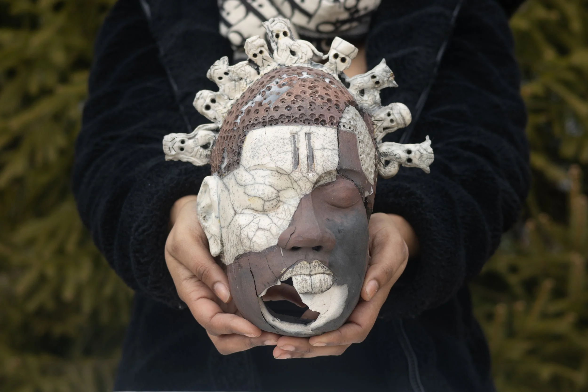 Person holding a decorative skull mask with ornate skull figures on top, made of ceramic or resin.
