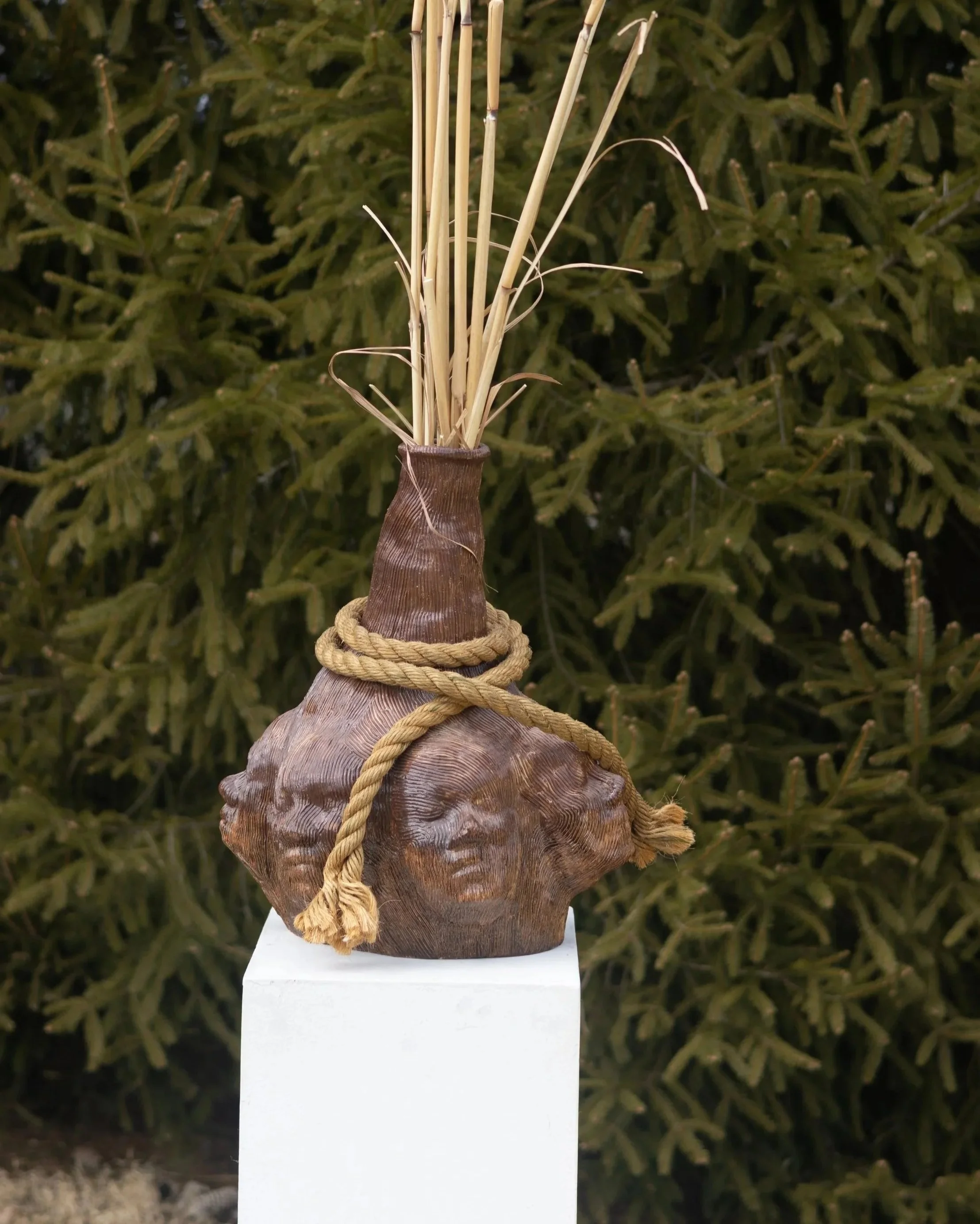 Traditional Japanese shōsō, a ceramic vessel wrapped with a thick rope, with several tall, thin wooden sticks bundled inside, displayed on a white pedestal with a green leafy background.