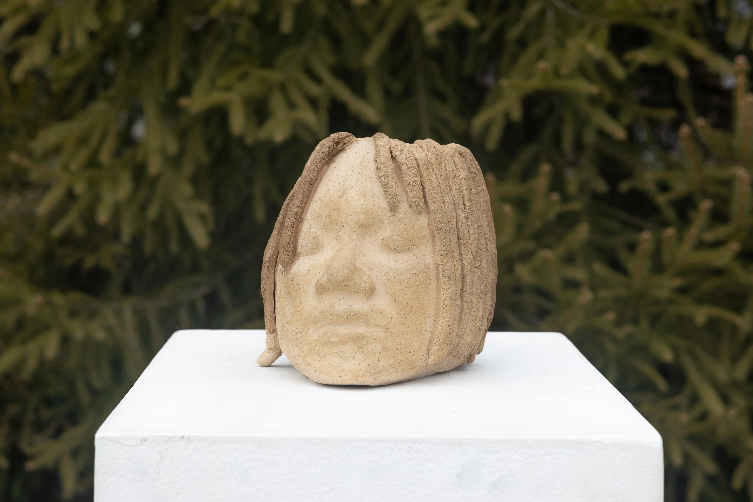 Sand sculpture of a face with detailed features on a white pedestal outdoors with greenery in the background.
