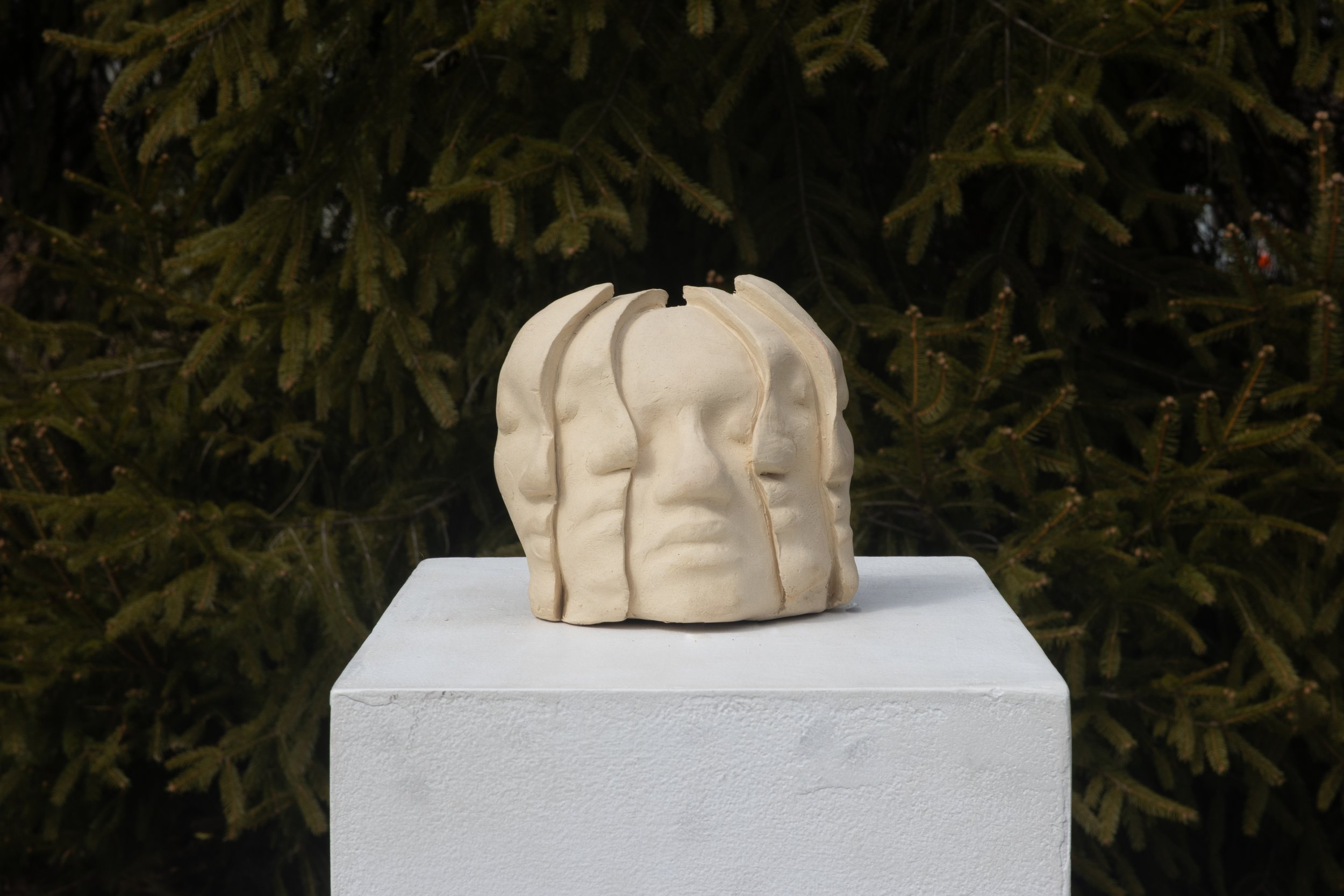 A white sculptural art piece resembling human heads in profile, arranged in a circular pattern, placed on a white pedestal outdoors with evergreen trees in the background.