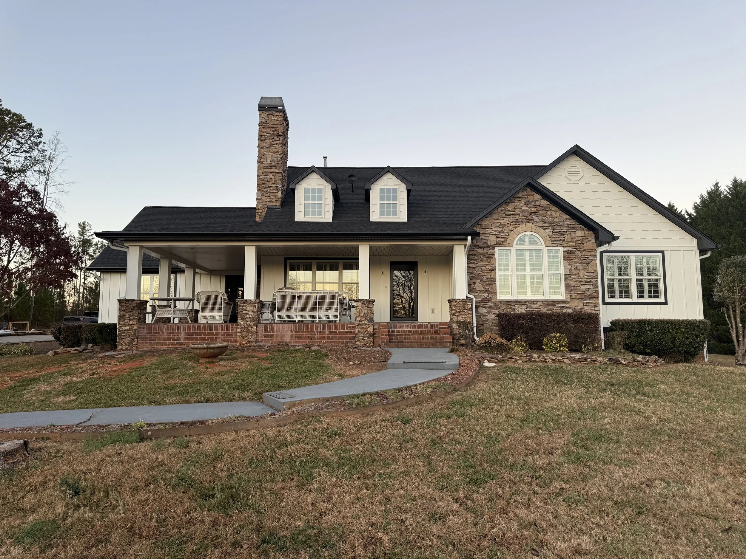 Front view of a two-story house with brick and white siding exterior, a chimney, a covered porch with outdoor furniture, and a landscaped yard with a curved pathway.