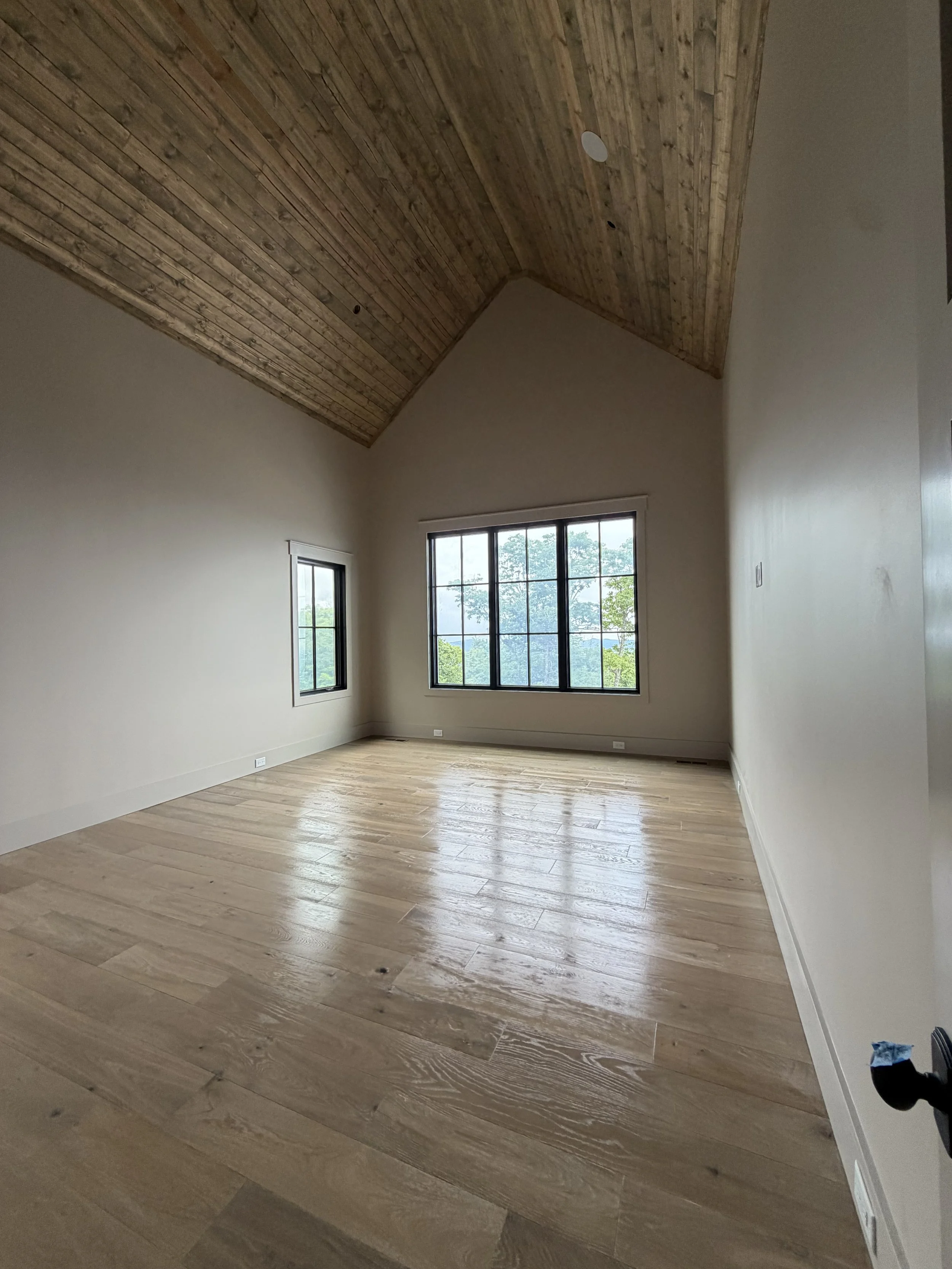 Empty room with light-colored wooden floor, white walls, large windows showing greenery outside, and a vaulted wooden ceiling.