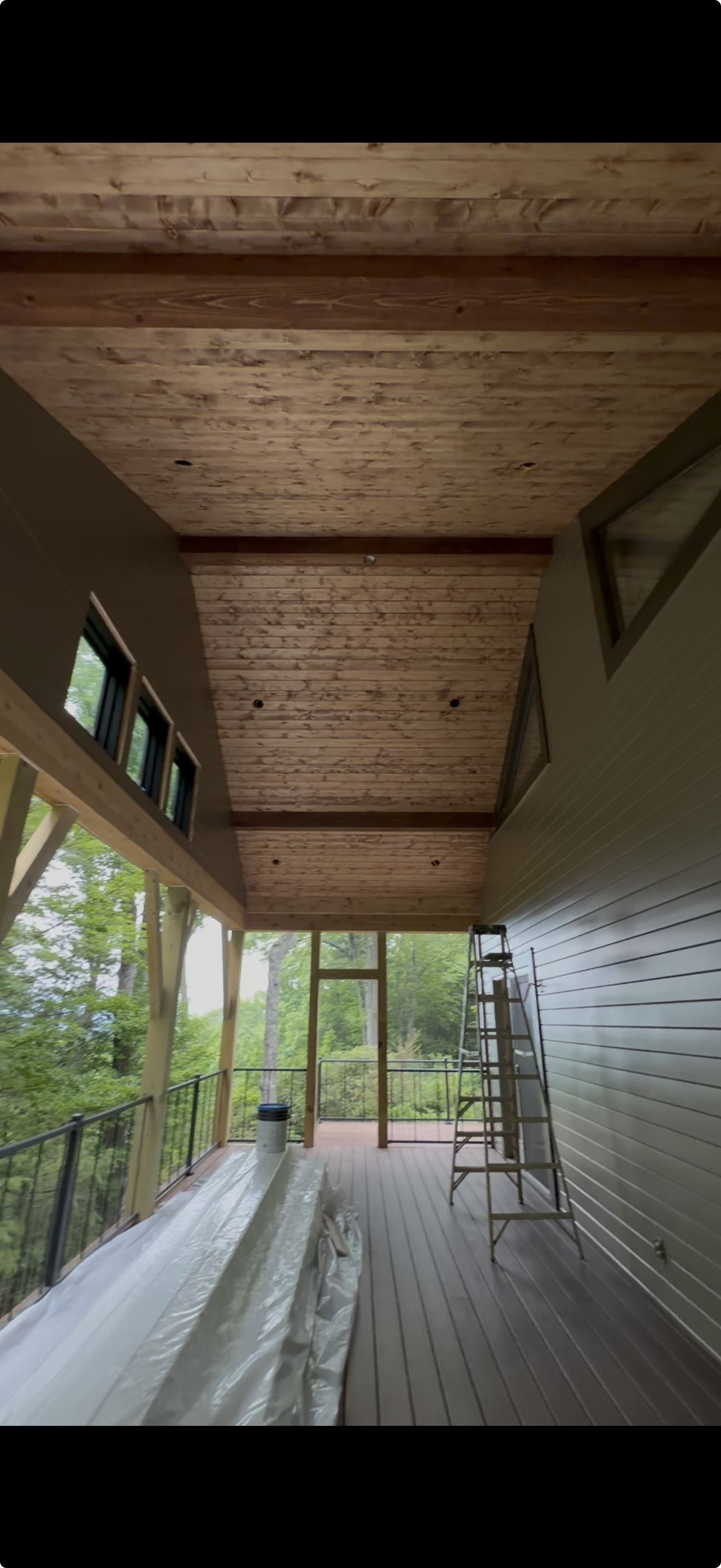 A covered outdoor porch or balcony with wood paneling on the ceiling, large windows, and a ladder. There are construction materials and a bucket on the floor, and the space overlooks a green, wooded area.