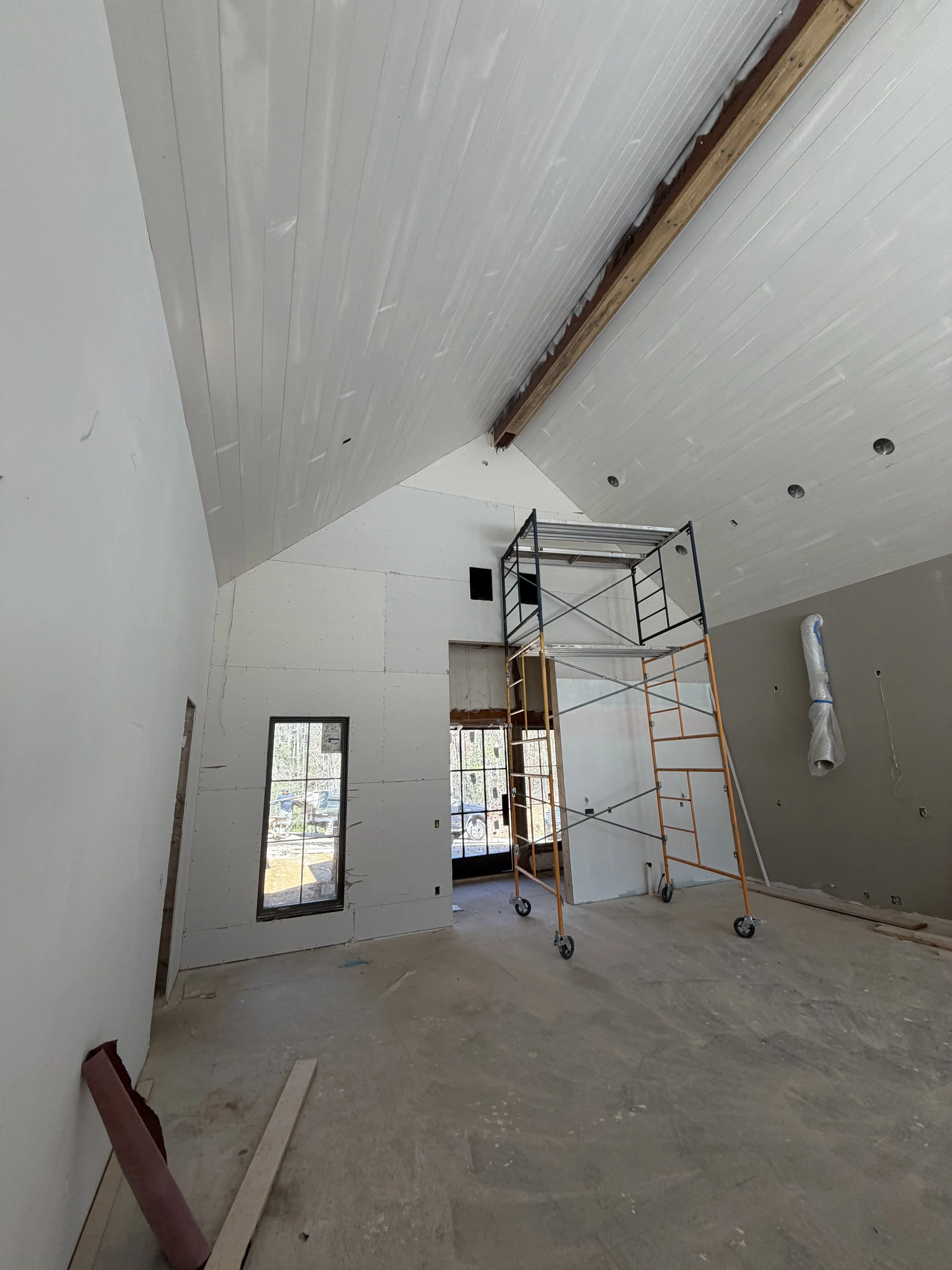 Interior of a room under construction with unfinished walls, a scaffold in the center, and windows allowing natural light to enter.