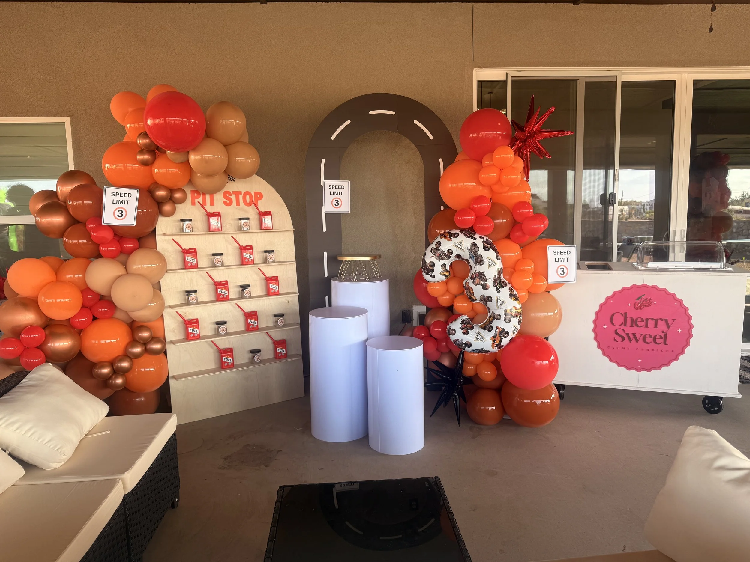 Decorative balloon arrangements in shades of orange, red, and beige, with a star-shaped balloon on the right, and a backdrop featuring signs reading 'Speed Limit 3' in a setting with glass doors and windows.