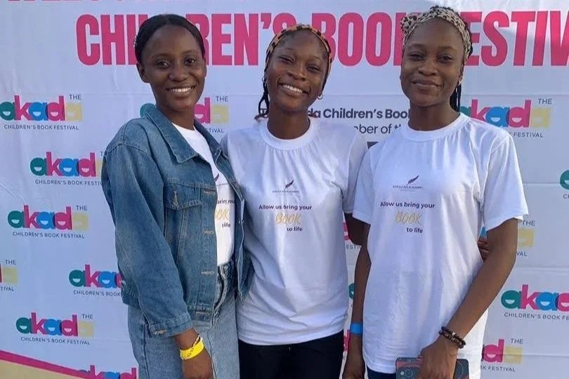 Three young women standing in front of a pink and white backdrop with the words 'Children's Book Festival' and 'OKAD' logo, smiling at the camera. Two of them are wearing white T-shirts with the slogan 'Allow us bring your book to life.' One woman is wearing a denim jacket.