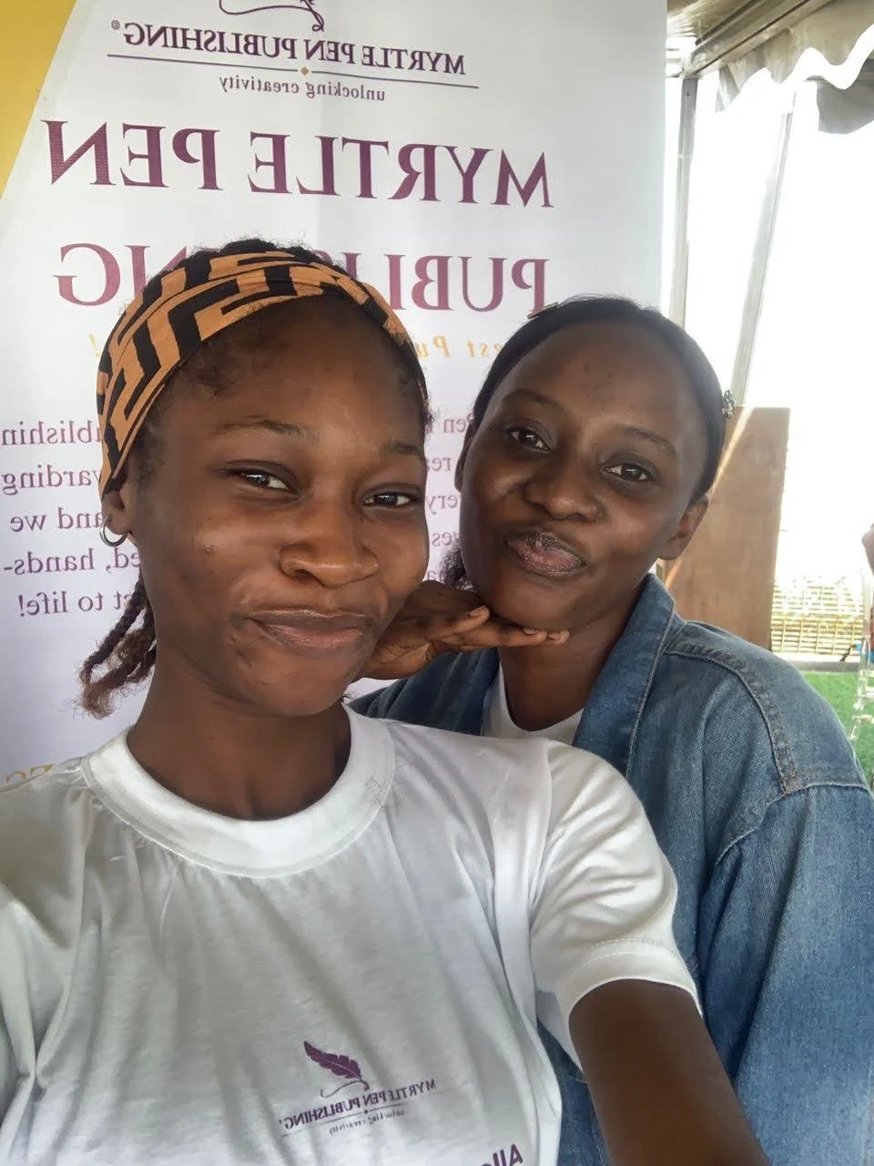 Two women taking a selfie together at an outdoor event, with a banner behind them that reads "Myrtle Pen Publishing" and some other text.