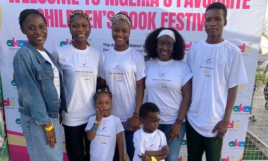 Group of seven smiling children and women standing in front of a pink and white banner at a children's book festival outdoor event.