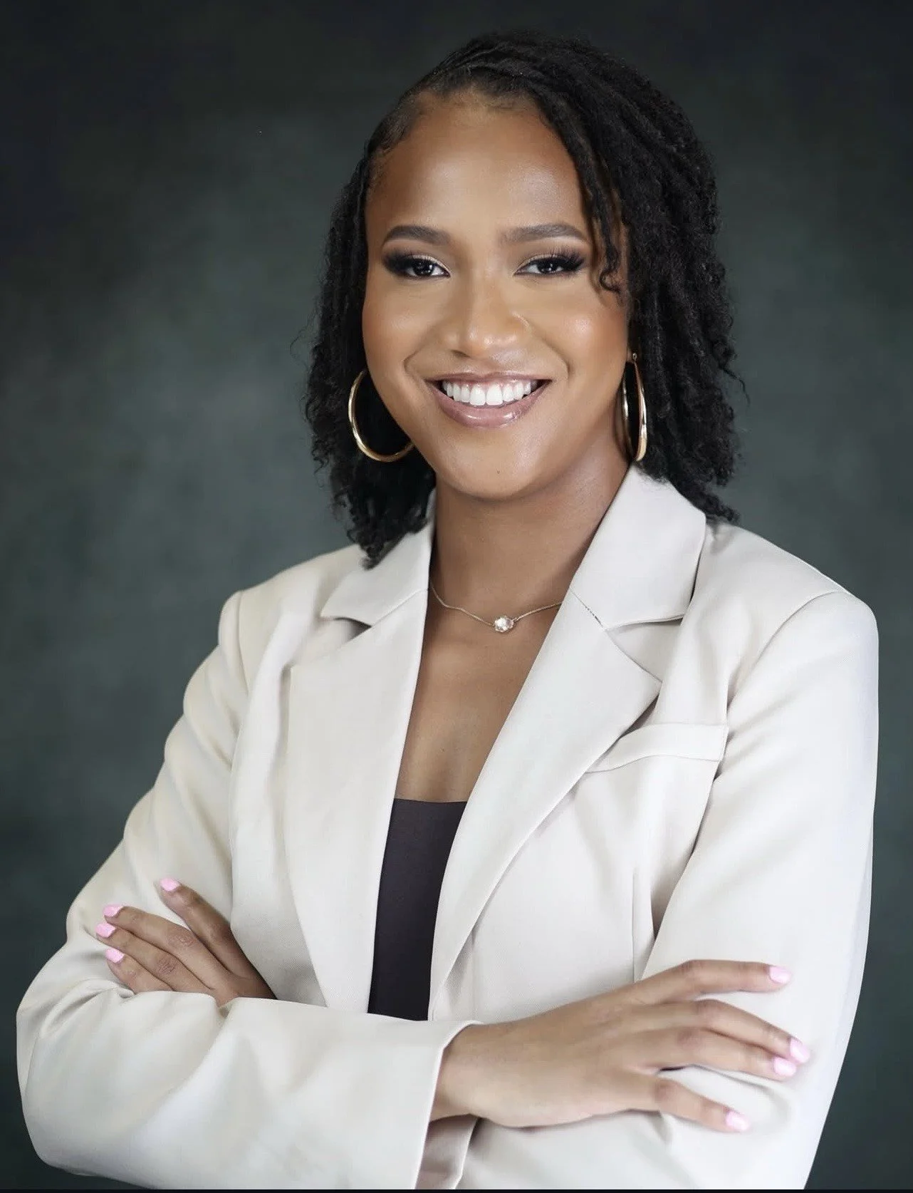 A smiling woman with dark curly hair, wearing hoop earrings, a necklace, a beige blazer, and a black top, standing with arms crossed against a dark clouded backdrop.