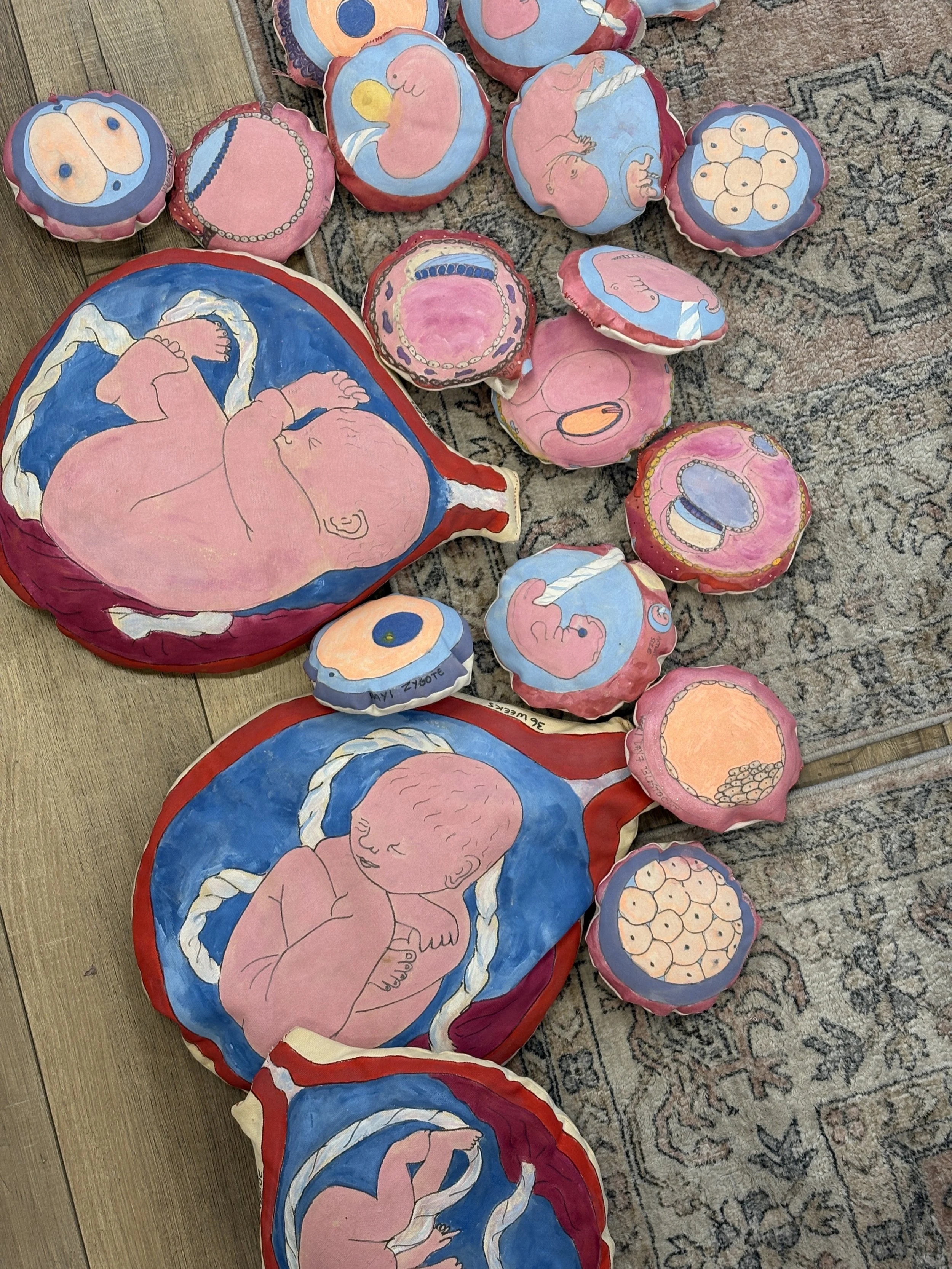 Colorful fabric cushions and pillows with hand-painted illustrations of babies and baby-related items, arranged on a patterned rug and wooden floor.