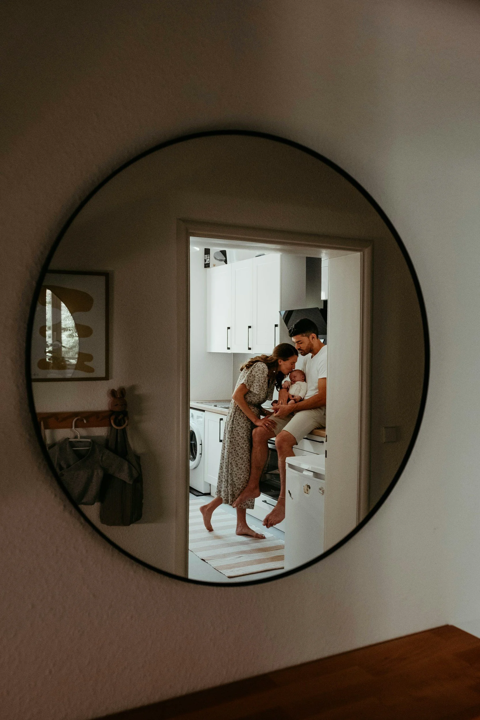 A family of three, a man, a woman, and a baby, are inside a kitchen, reflected in a round mirror mounted on the wall.