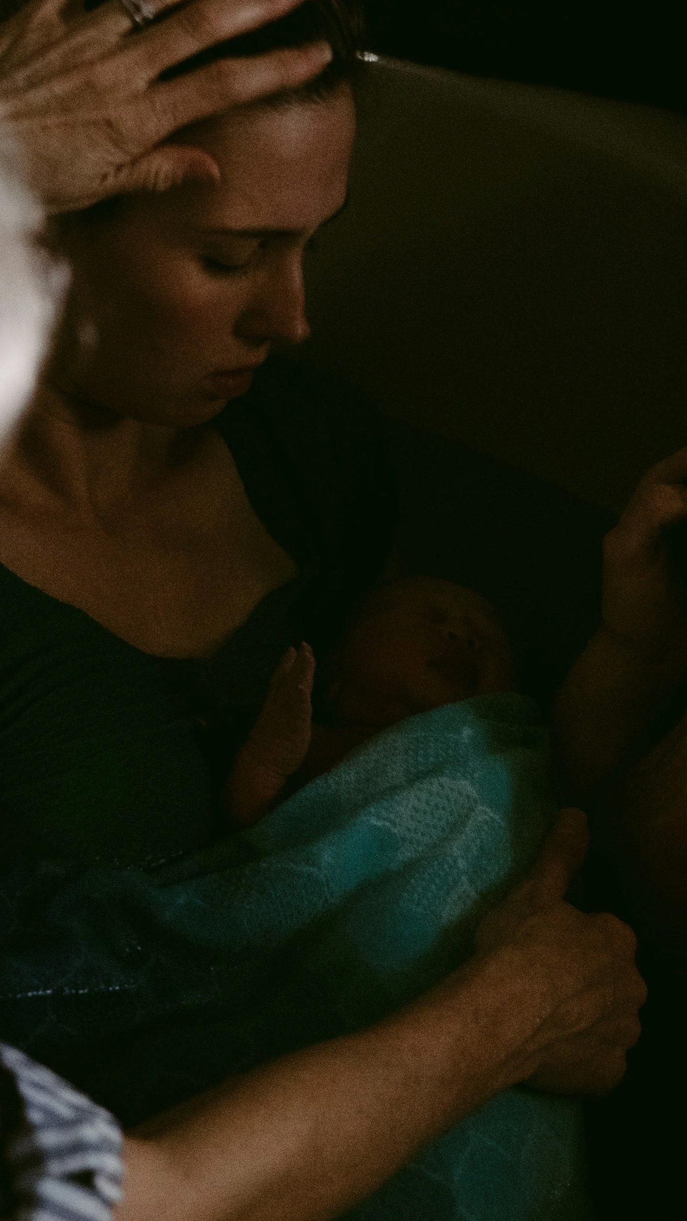 A woman with closed eyes and a serious expression is holding a sleep baby with a pacifier, wrapped in a blanket, lying in her arms in a dark environment.