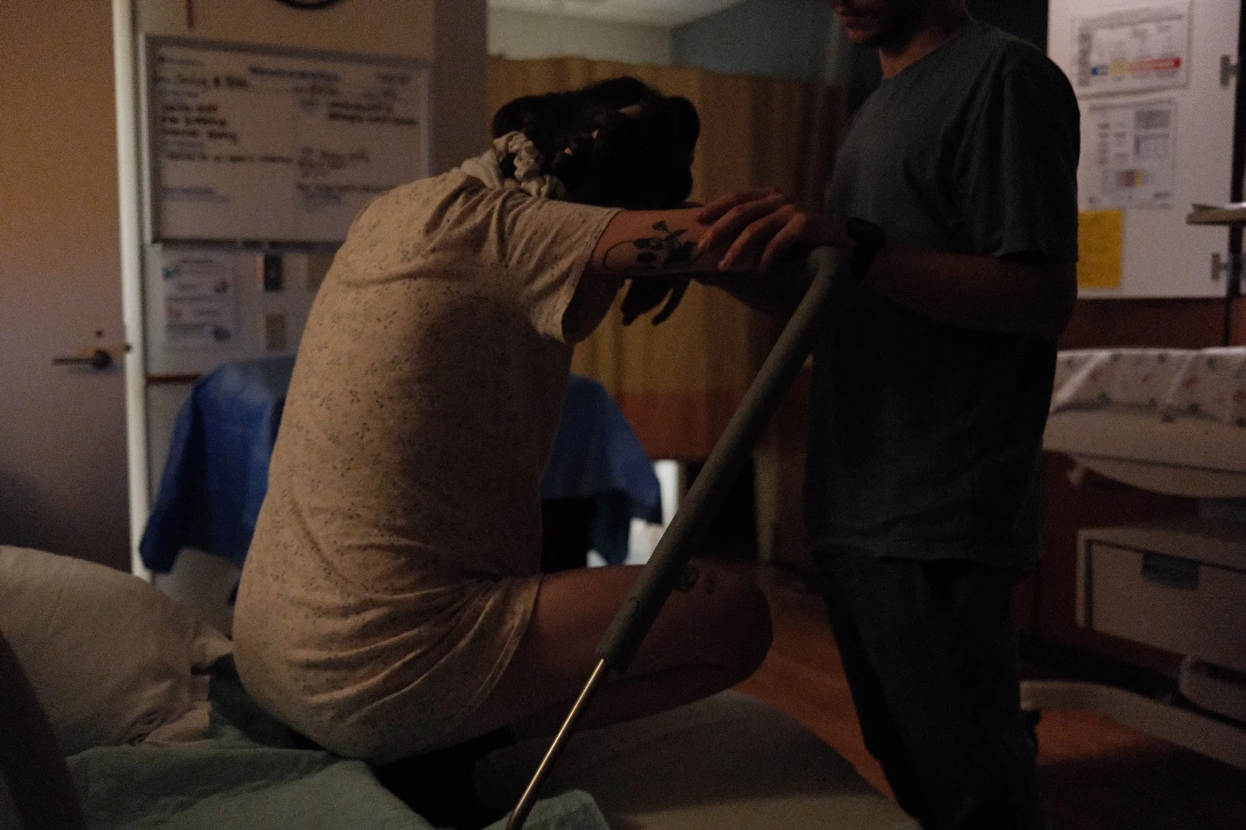 A person with dark hair and a tattoo on their arm, wearing a beige speckled shirt, leaning over to hold the hand of a man sitting or lying on a hospital bed. The man in bed has dark curly hair and is wearing a light-colored shirt. The setting appears to be a hospital room with medical posters on the wall.
