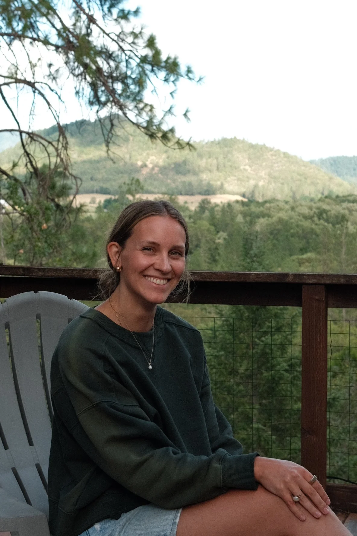 Smiling woman sitting outdoors on a deck with mountain and forest background during daytime.