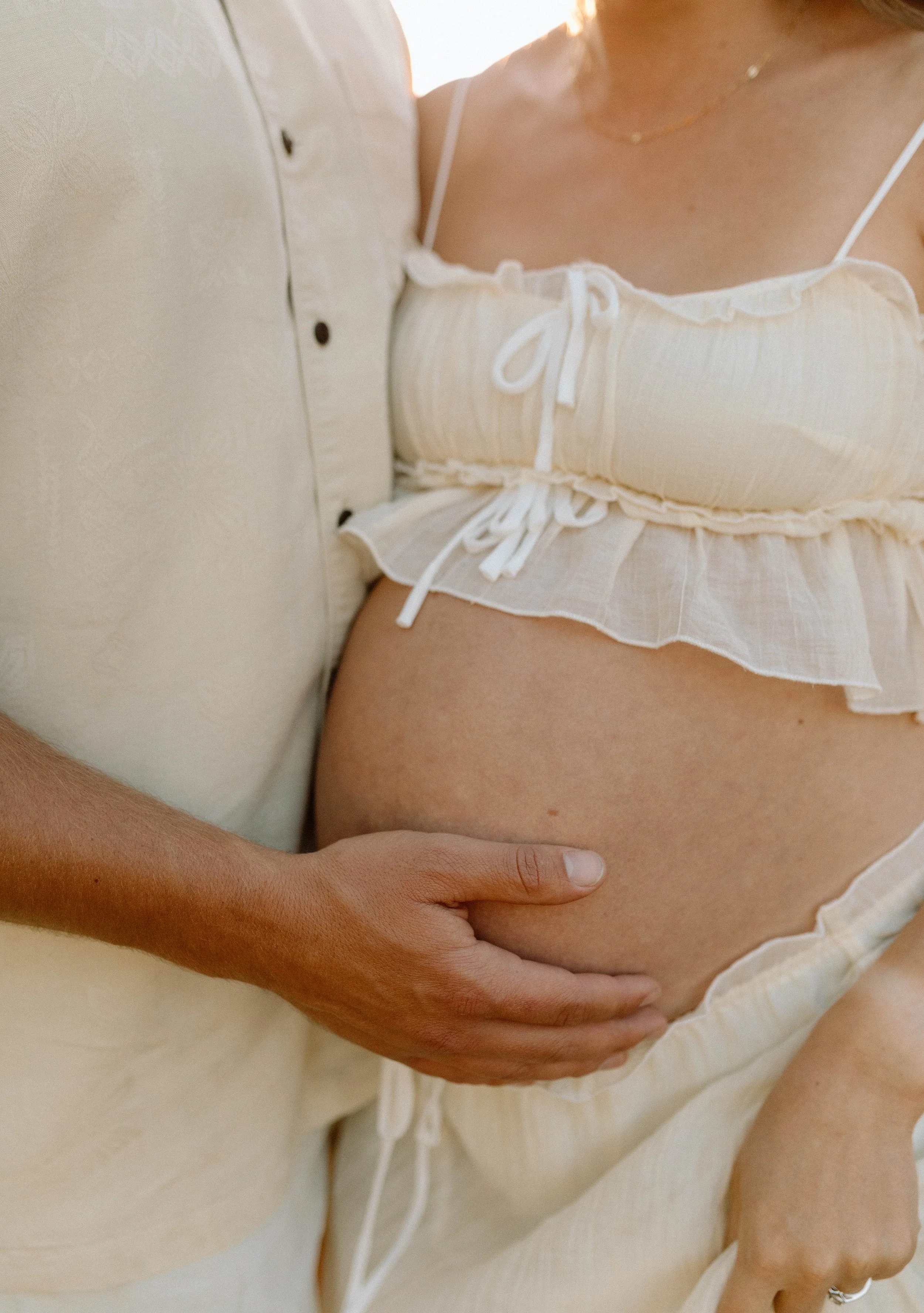 Close-up of a woman in a beige, frilly, spaghetti-strap top, with a hand gently placed on her waist. The background is a soft, warm light.
