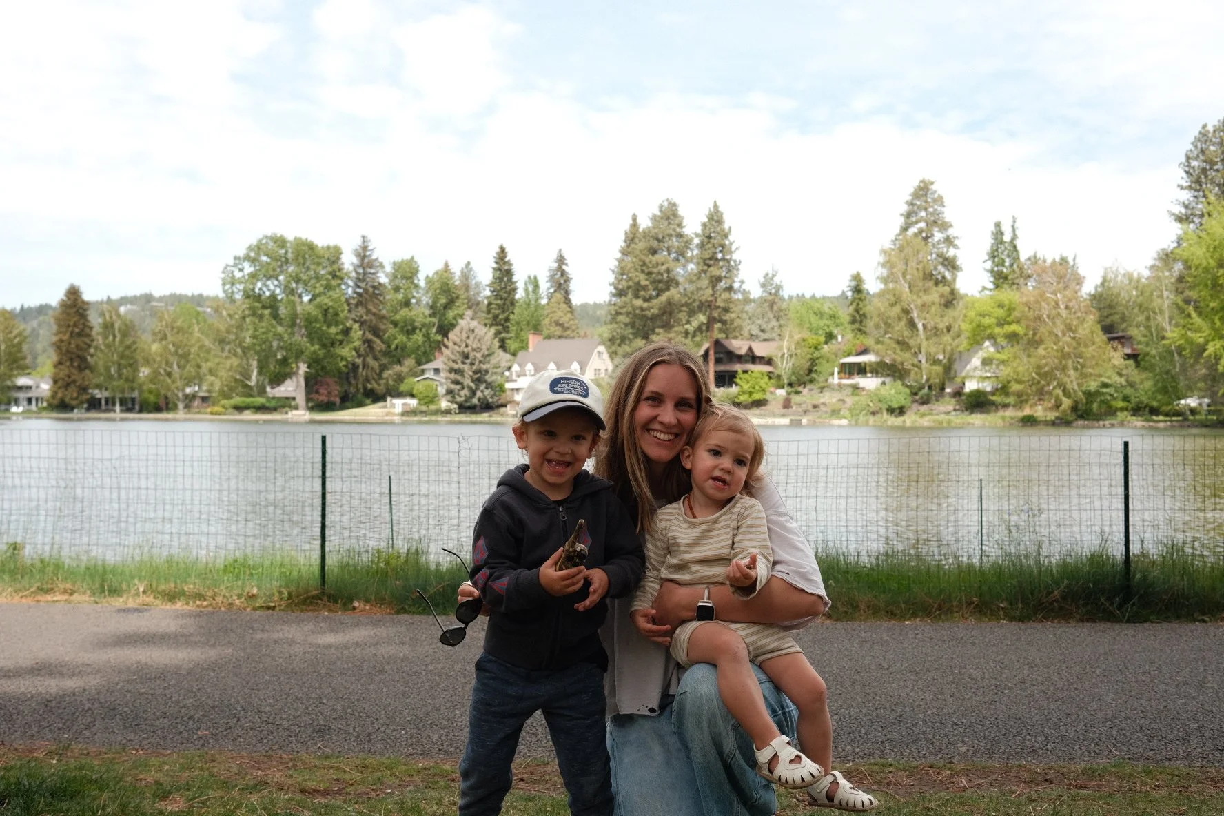 A woman with two young children sitting by a lake with houses and trees in the background, smiling for the camera.