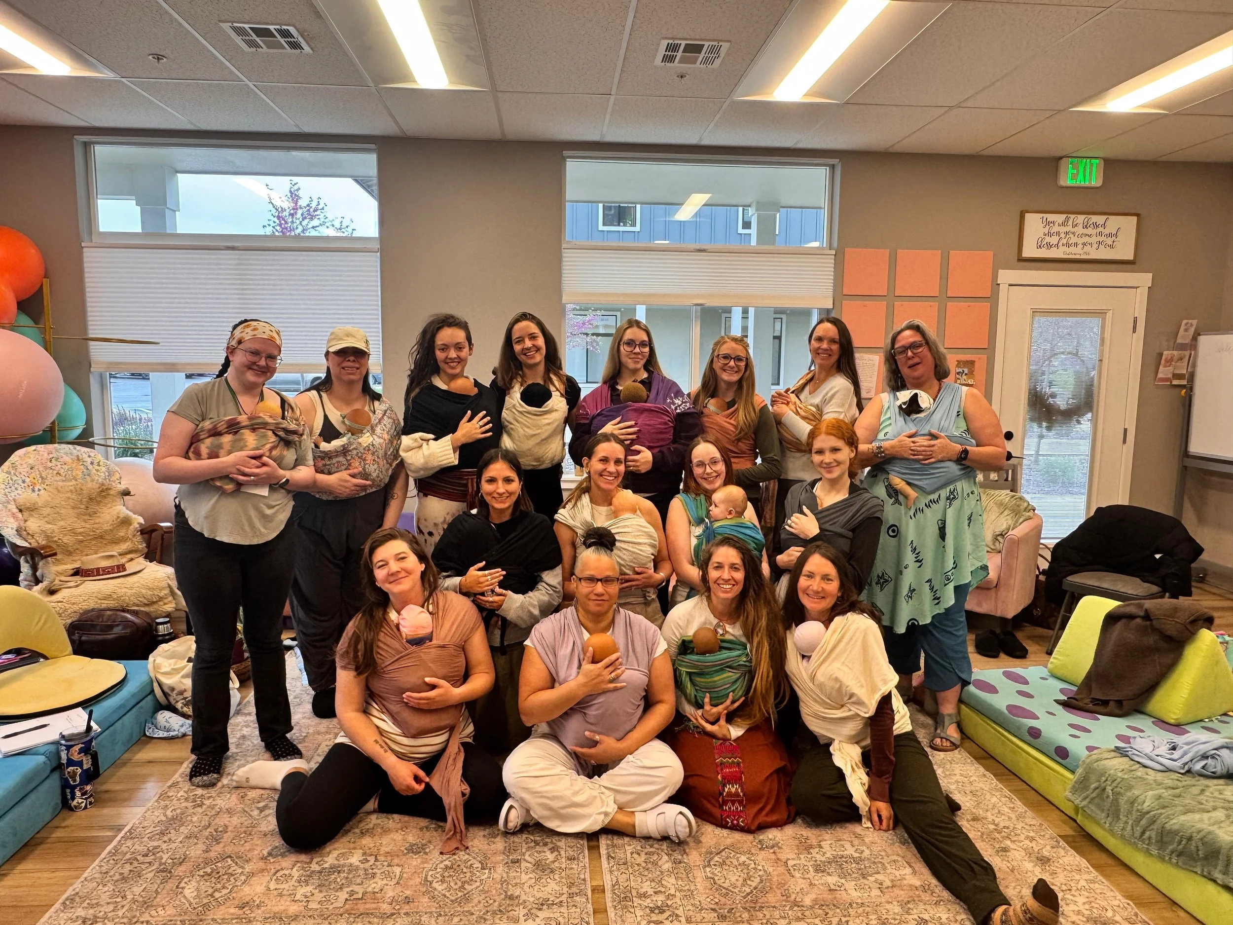 Group of women and children gathered indoors, some holding babies, standing and sitting on a patterned rug in a bright room with large windows, colorful furniture, and wall decorations.