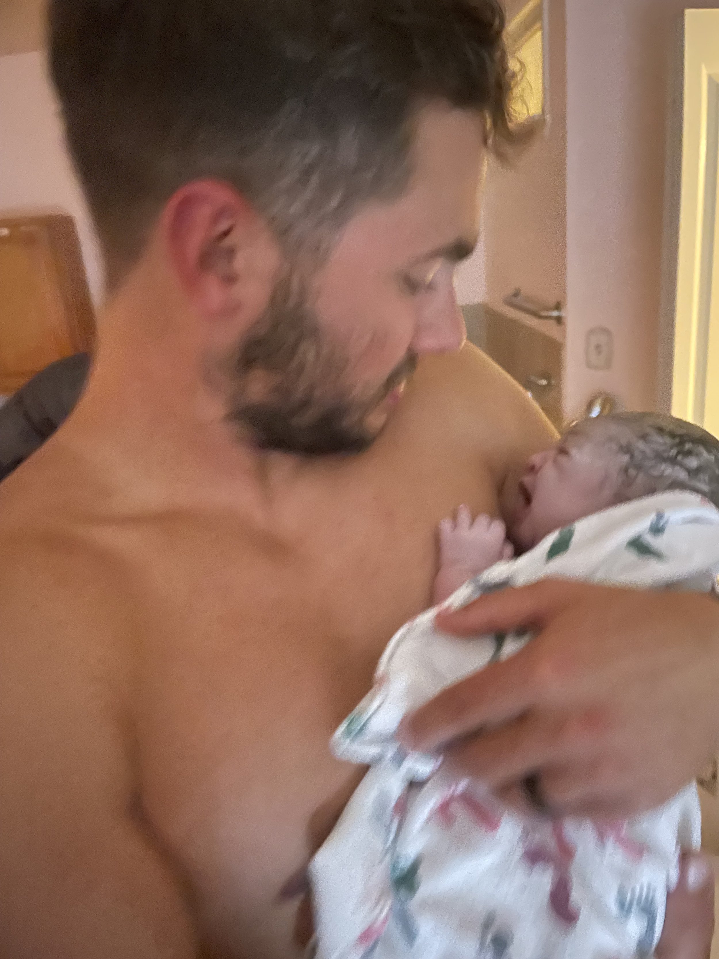 A man holding a newborn baby, close-up of their faces with the man smiling and looking at the baby's face, in a hospital or home setting.