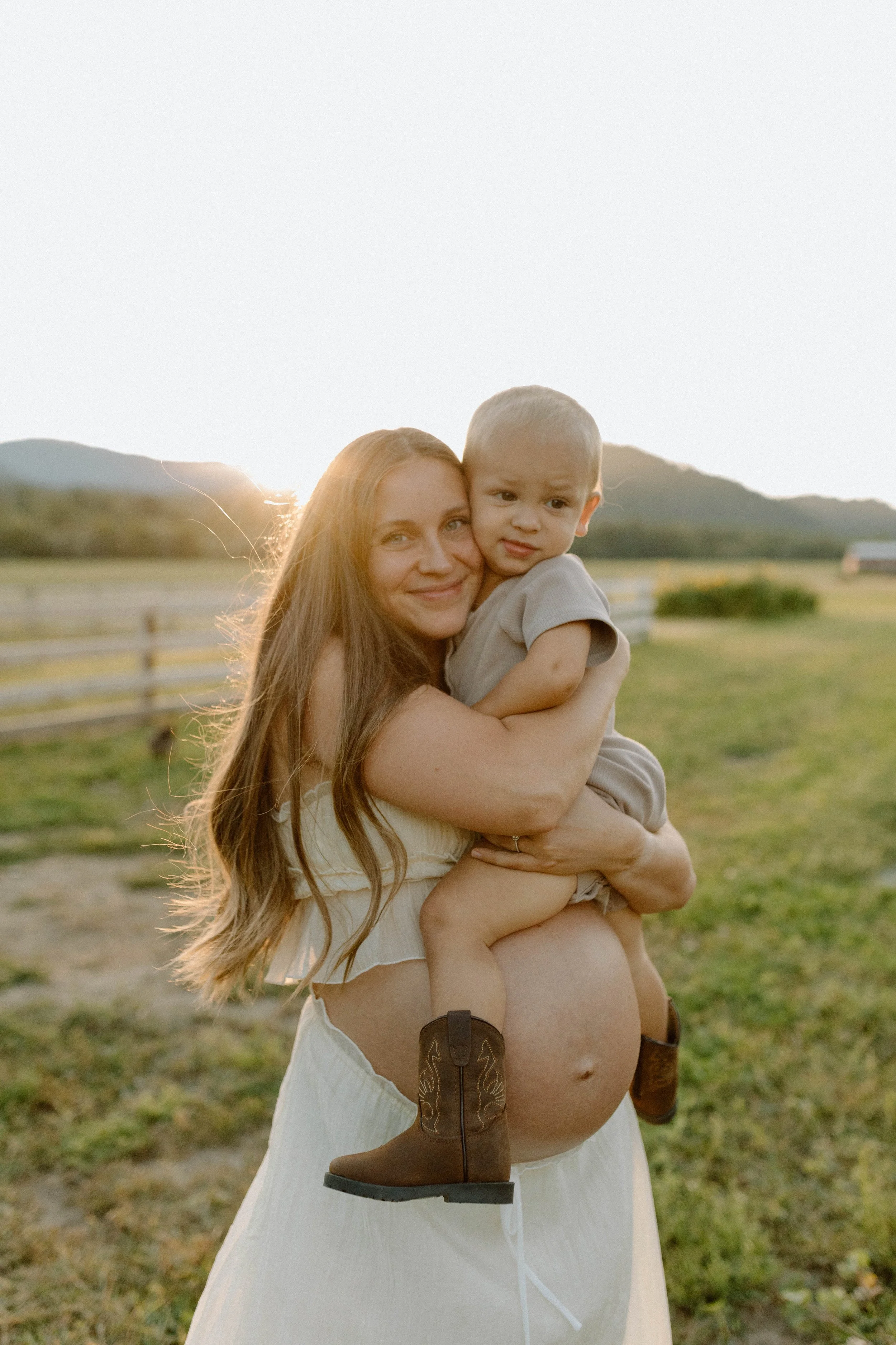 A woman with long brown hair holding a young boy in a rural outdoor setting during sunset. The boy is wearing cowboy boots and a beige shirt, looking to the side with a curious expression. The background features green fields, a wooden fence, and mountains.