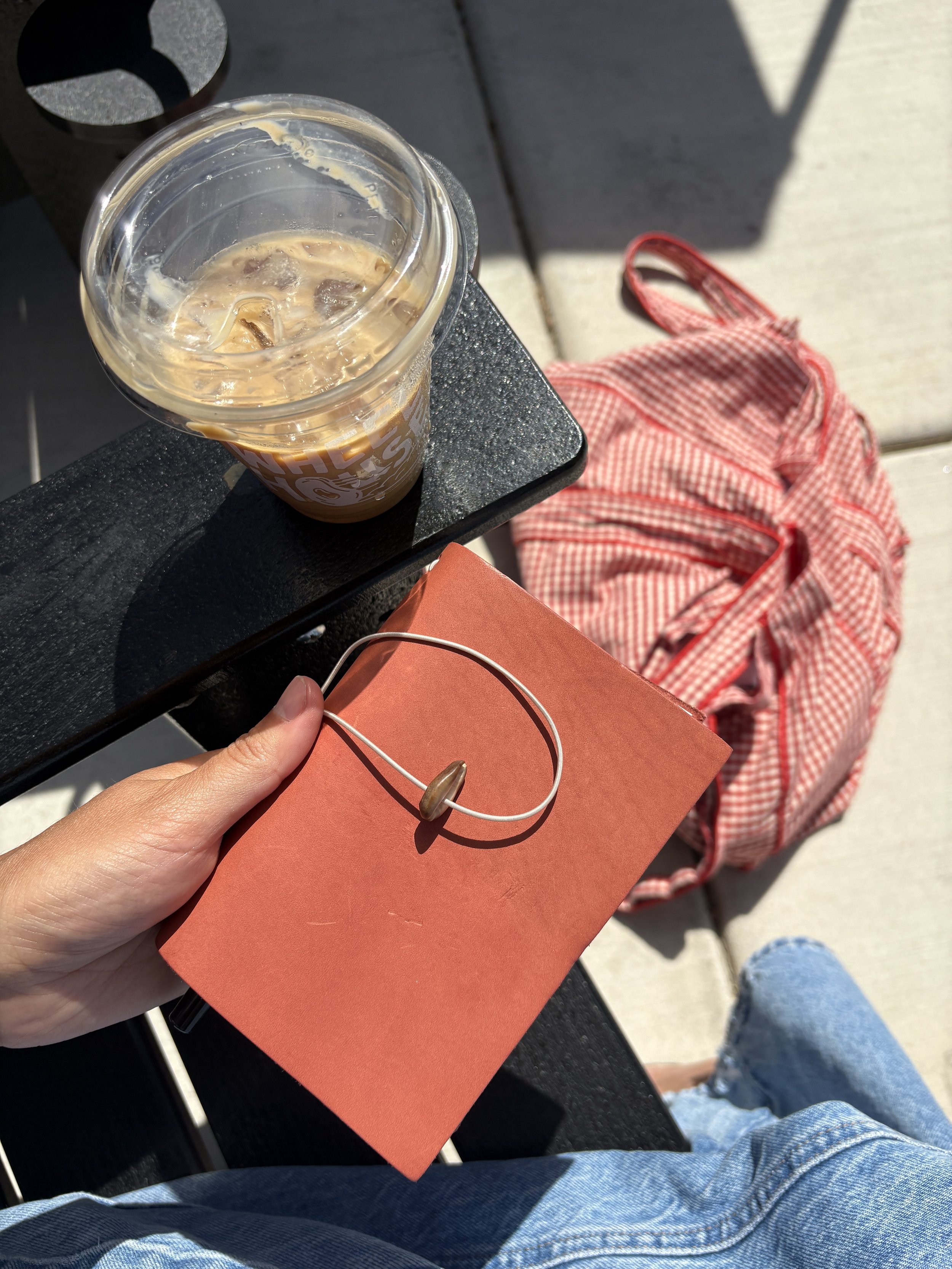 Top view of an iced coffee cup on a black table, a person's hand holding a red bag, and a red checkered backpack on the sidewalk with a person's legs in jeans visible.