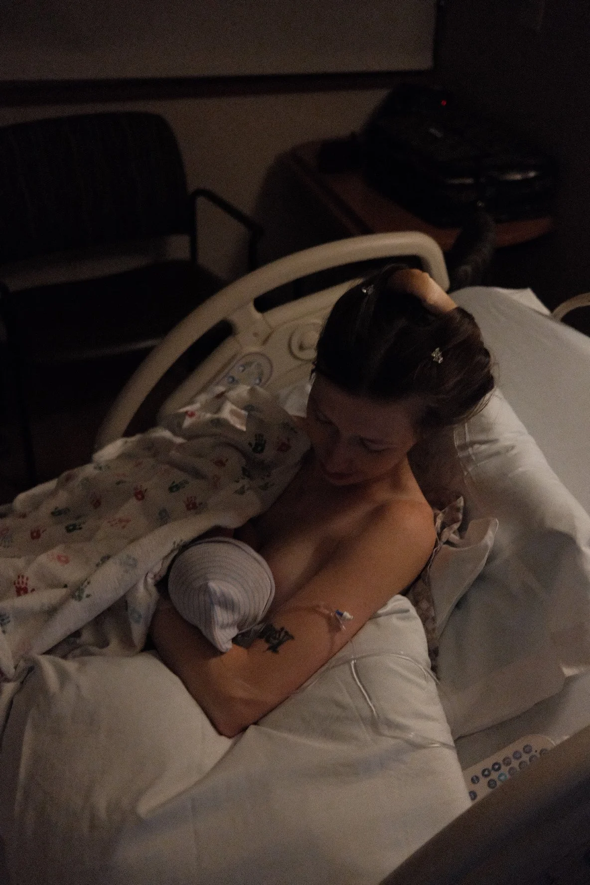 A woman in a hospital bed after giving birth, holding her newborn baby wrapped in a blanket. The room is dimly lit.