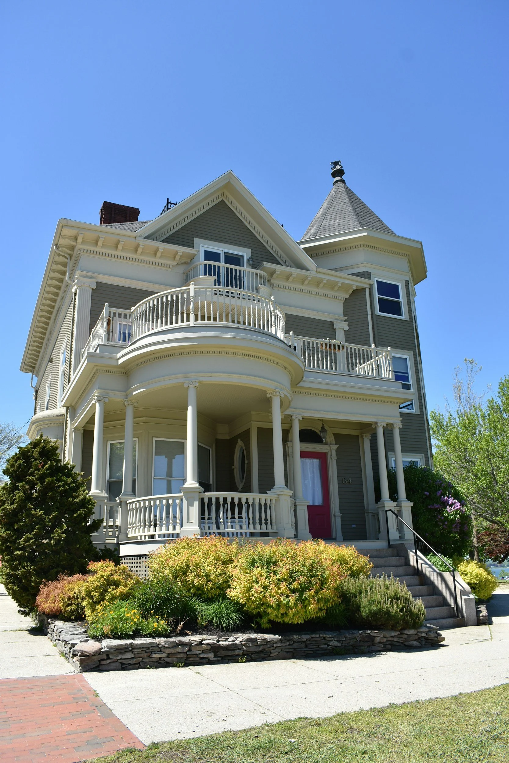 A large Victorian-style house with a cream-colored exterior, featuring a rounded front porch with white columns, a red front door, and a turret with a conical roof. There are landscaped bushes and flowers in the front yard, with a stairway leading up to the porch and a sidewalk in front.