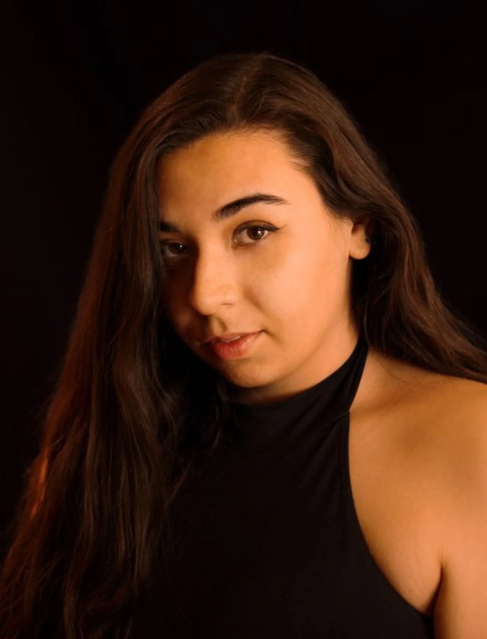 Close-up of a woman with long brown hair, smiling, wearing a black top, standing against a black background.