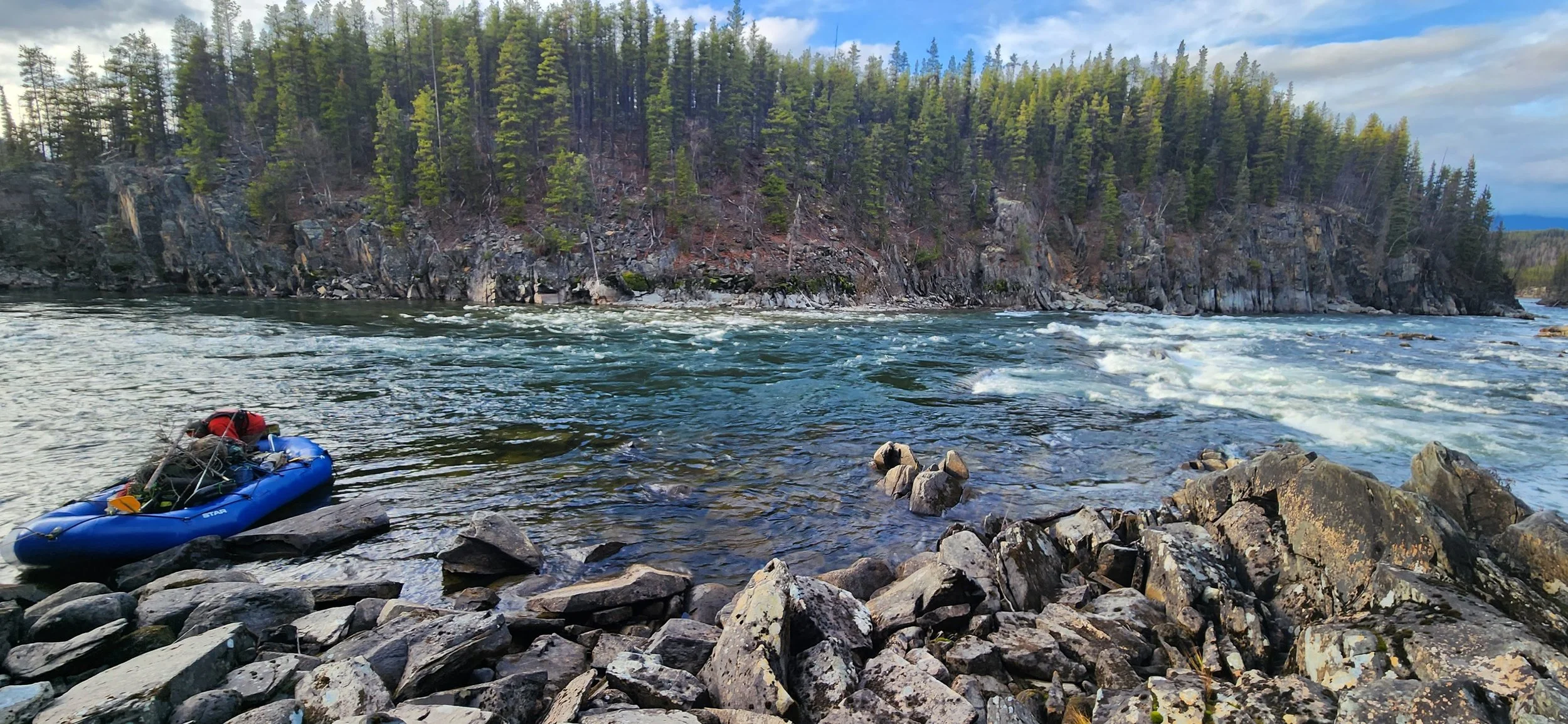 A river flowing through a rocky shoreline with a forested cliff in the background. A blue inflatable raft with gear is on the rocks near the water.