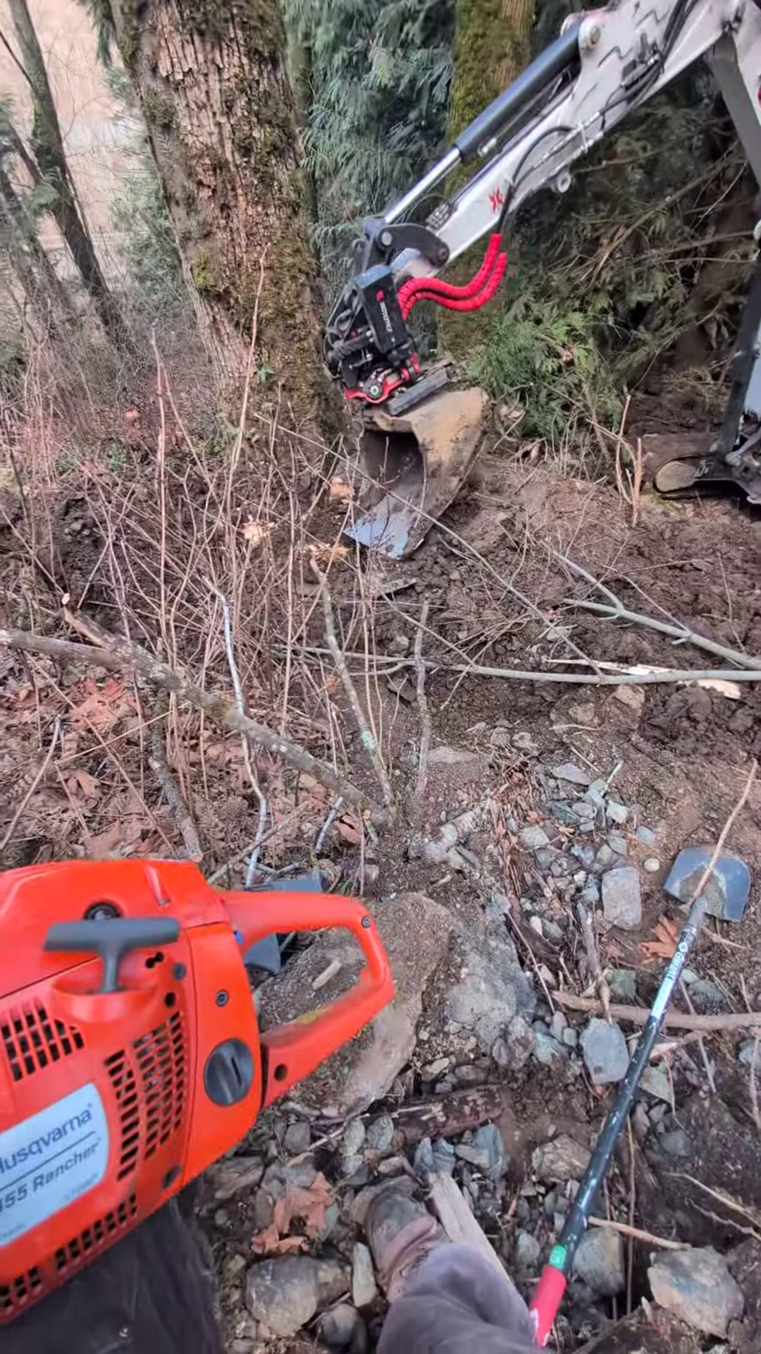 A person operating a chainsaw and an excavator with a hydraulic hammer attachment, road building in a forested area.