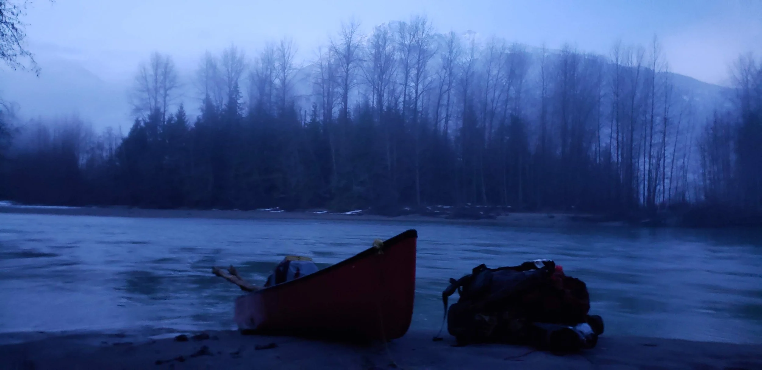 A river scene at dusk with a boat and backpacks on the shore, and a background of leafless trees and foggy mountains.